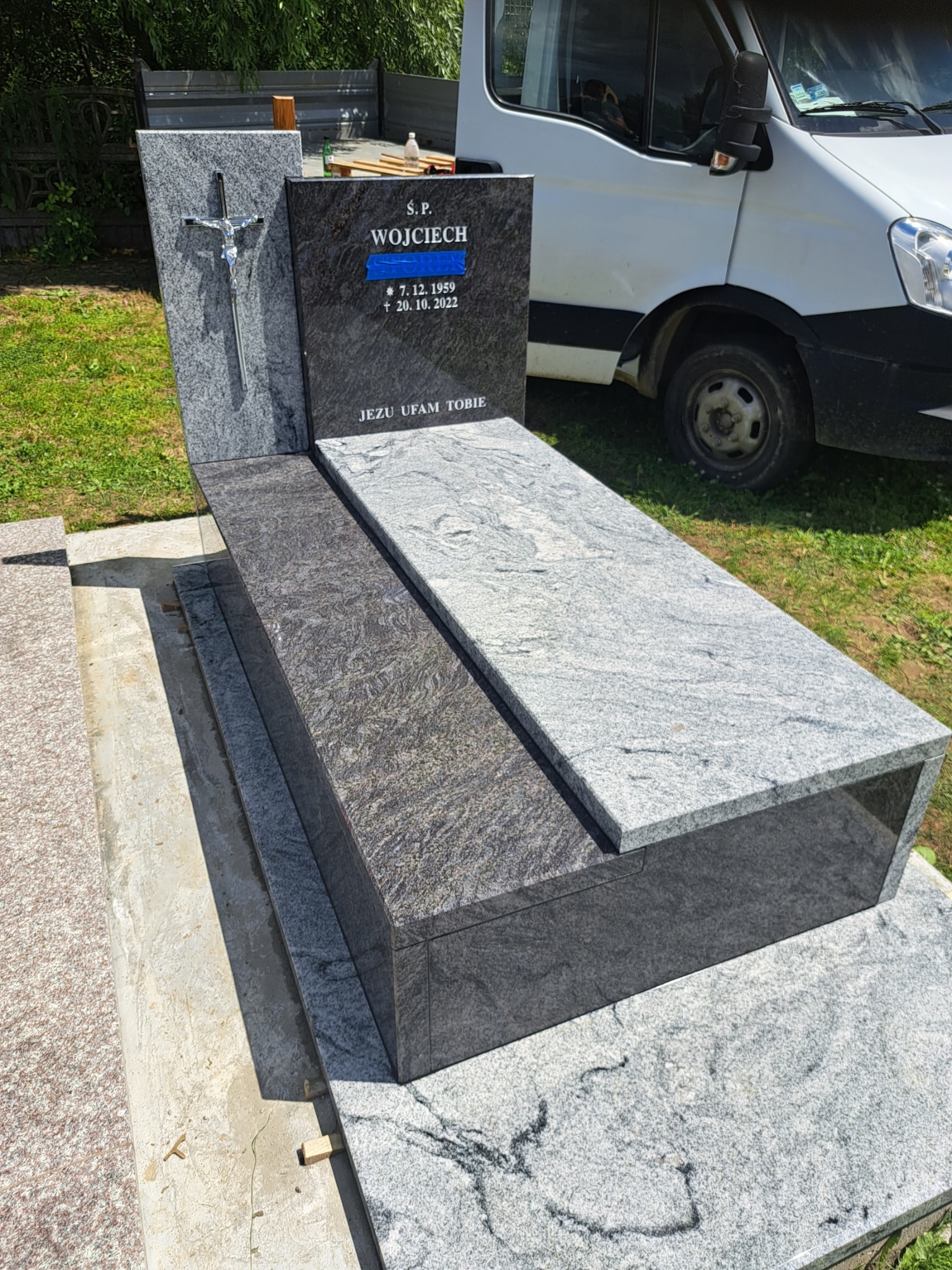 Granitowy nagrobek z krzyżem i inskrypcją 'Ś.P. Wojciech' z datami, umieszczony na cmentarzu, z białym samochodem dostawczym w tle.