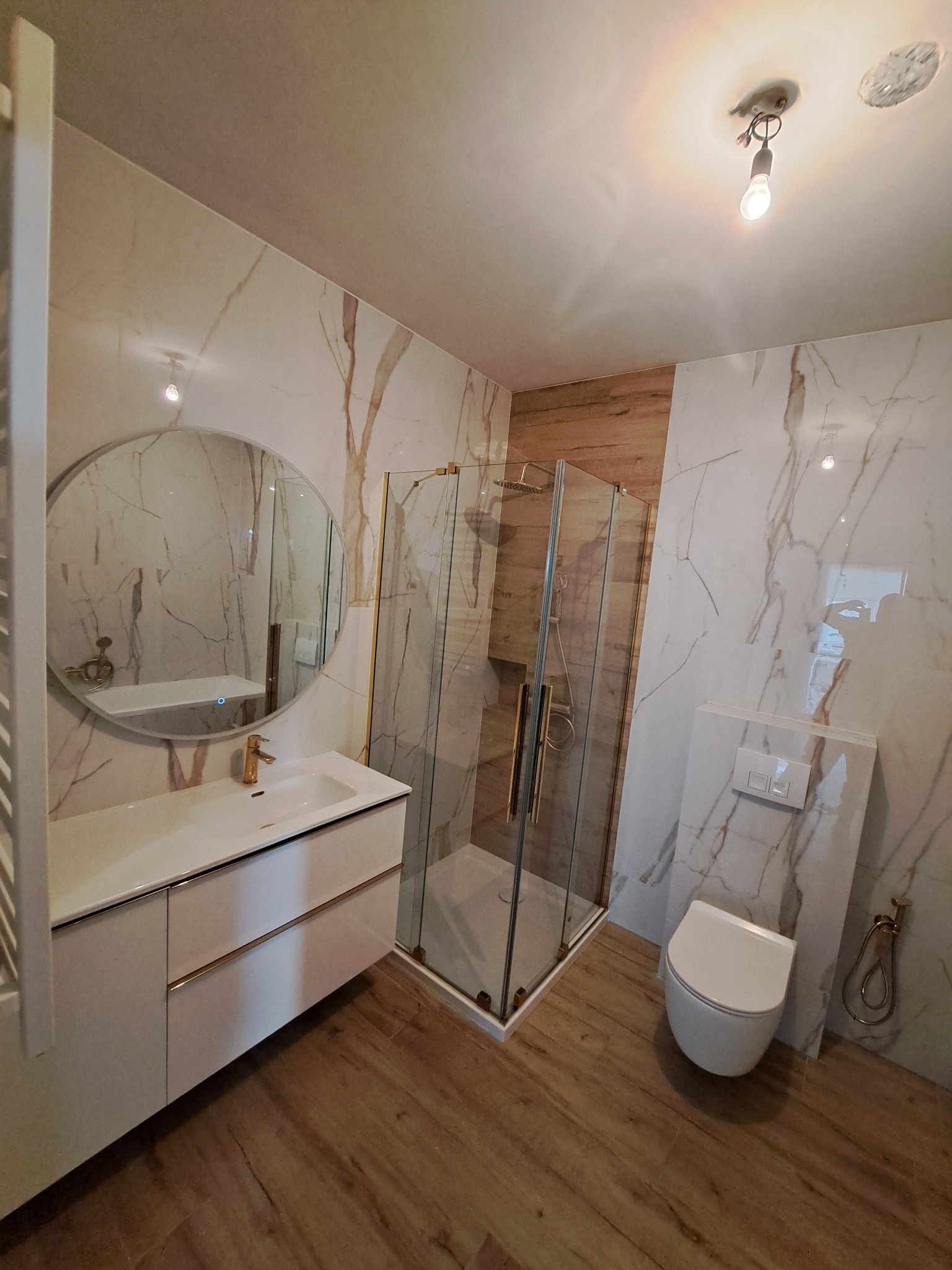 Nowoczesna łazienka z kabiną prysznicową, umywalką z szafką, okrągłym lustrem z podświetleniem i toaletą, wykończona płytkami imitującymi marmur i drewno.