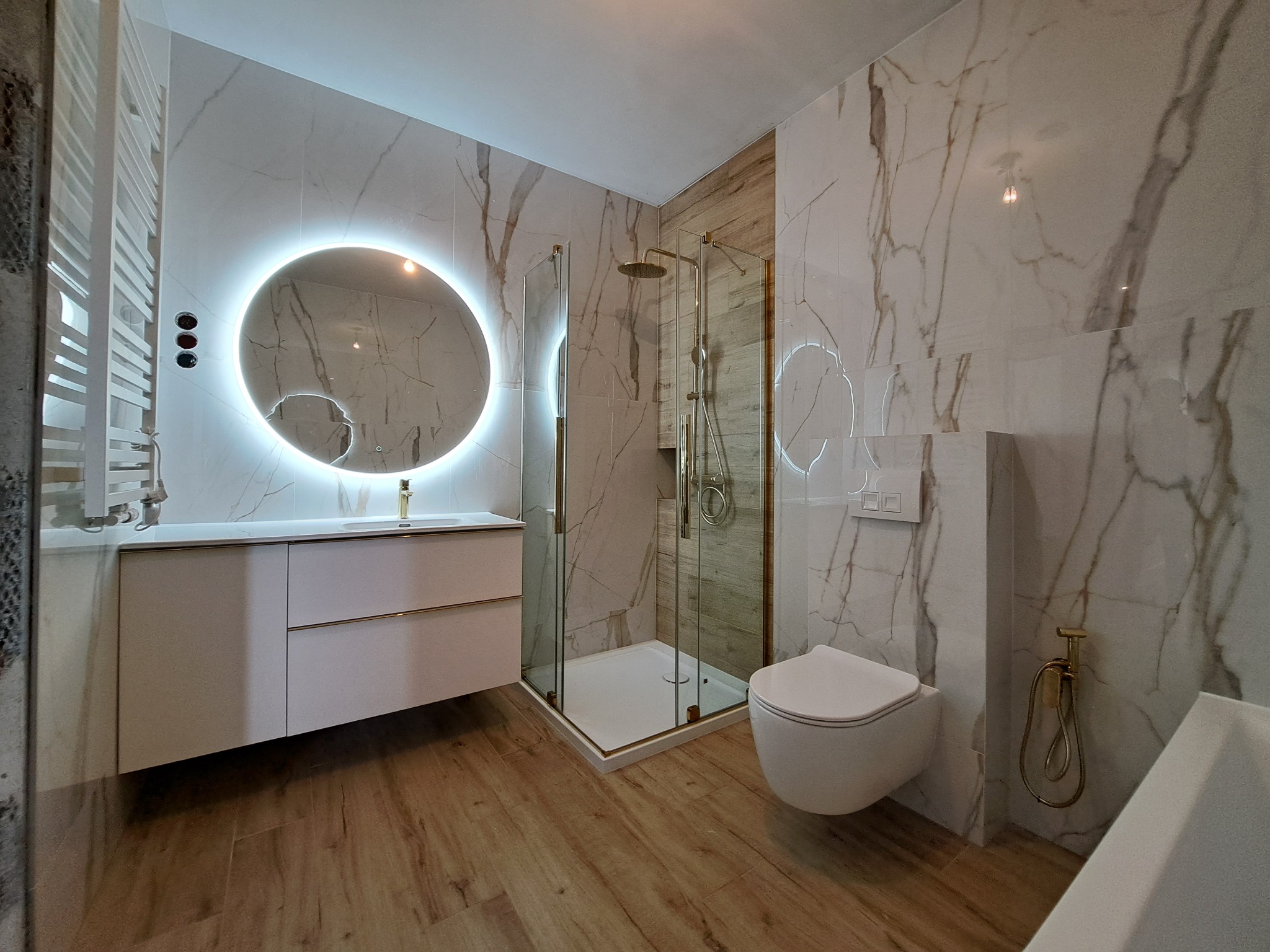 Nowoczesna łazienka z kabiną prysznicową ze złotymi detalami, umywalką z szafką, okrągłym lustrem z podświetleniem LED i drewnopodobną podłogą.
