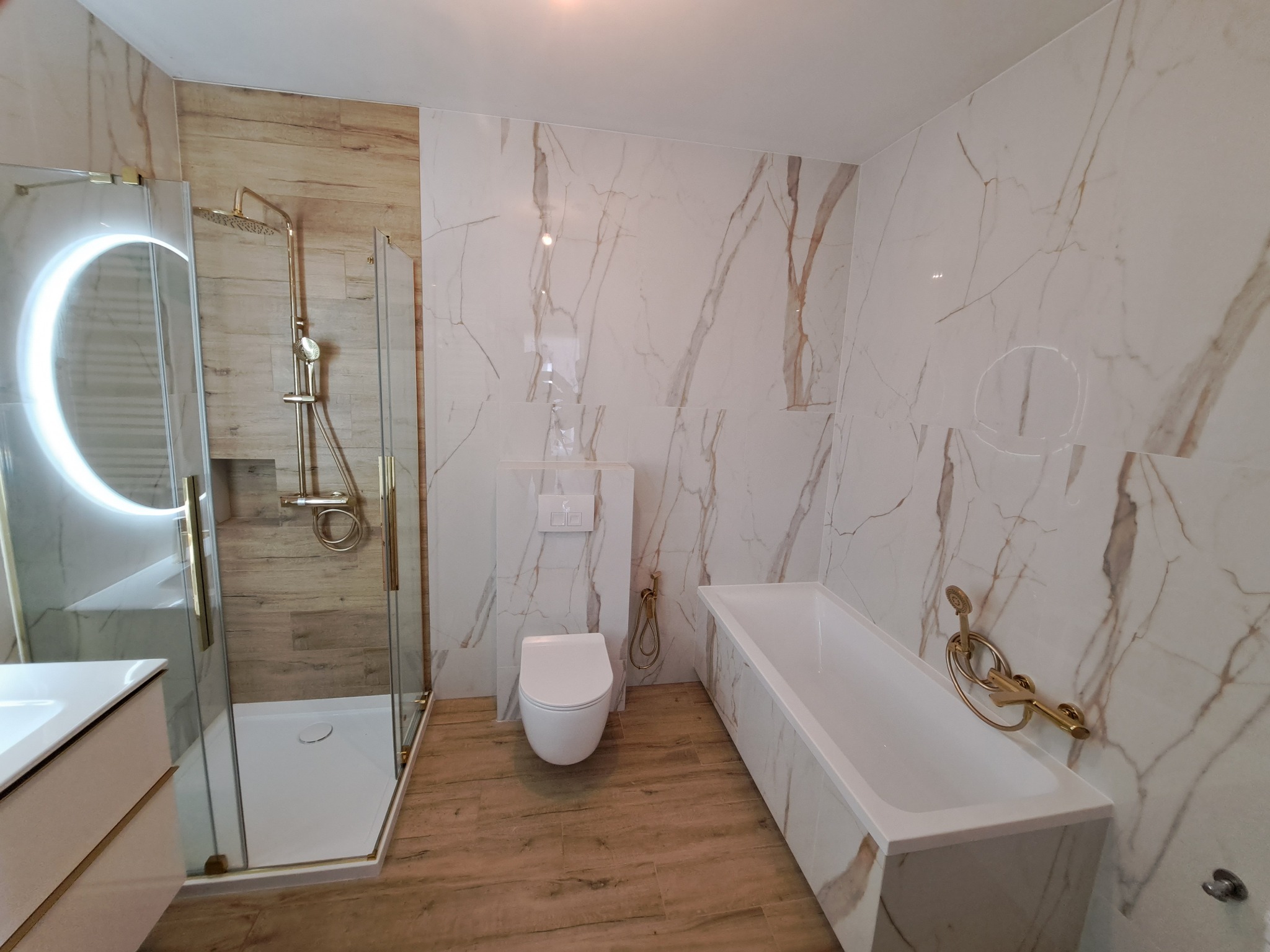 Nowoczesna łazienka z marmurowymi płytkami, złotą armaturą i drewnopodobną podłogą, widok na kabinę prysznicową z okrągłym lustrem LED, toaletę i wannę.
