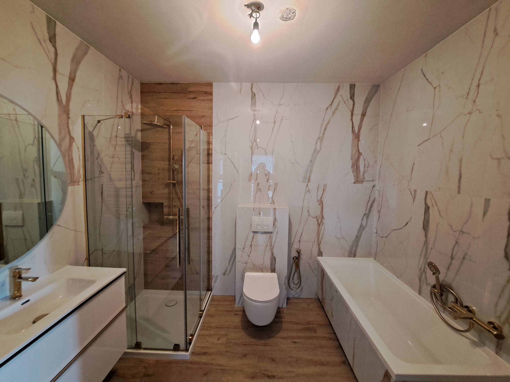 Wykończona łazienka z kabiną prysznicową, podwieszaną toaletą, umywalką z szafką i wanną, wyłożona płytkami imitującymi biały marmur z brązowymi żyłami oraz płytkami imitującymi drewno.