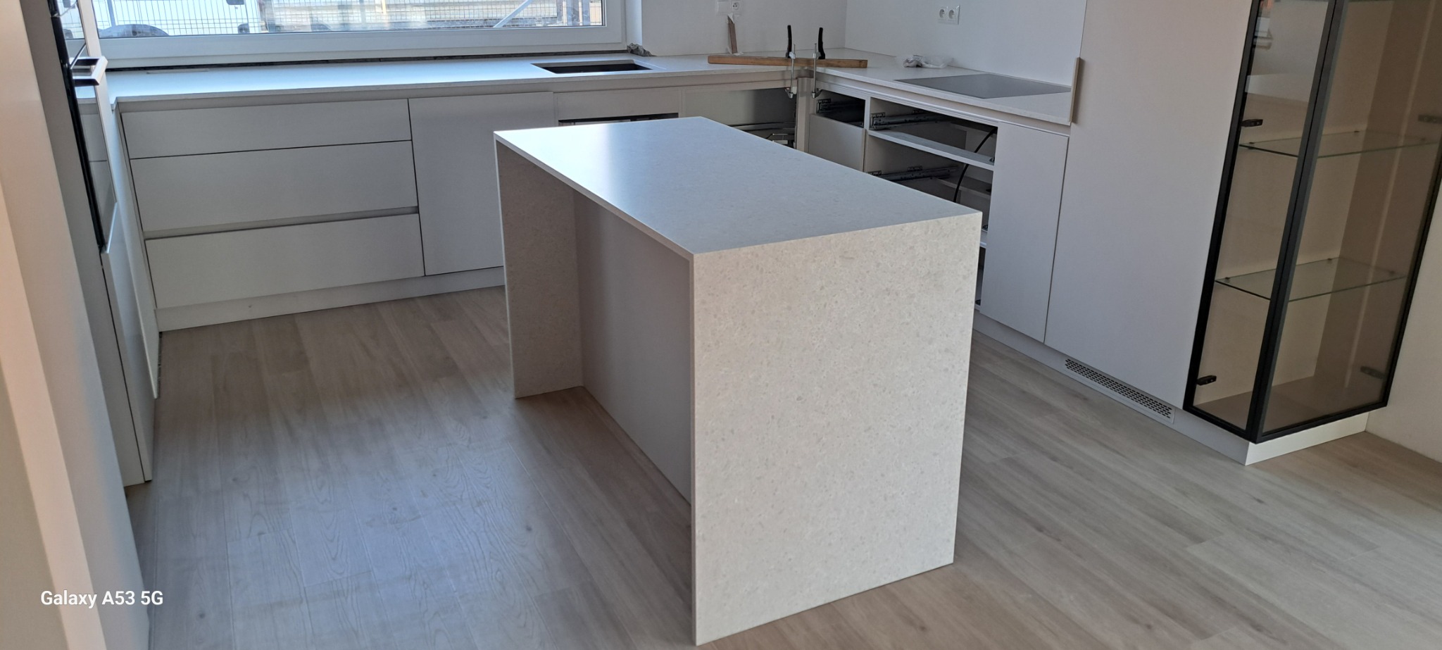 Nowoczesna, jasna kuchnia w trakcie montażu, z wyspą wykończoną jasnym konglomeratem, widoczne szuflady bez frontów i elementy montażowe.