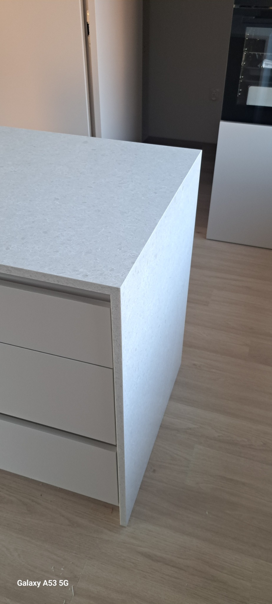 Szary blat z konglomeratu na wyspie kuchennej z szufladami, widok z bliska, z widoczną strukturą materiału i fragmentem podłogi.