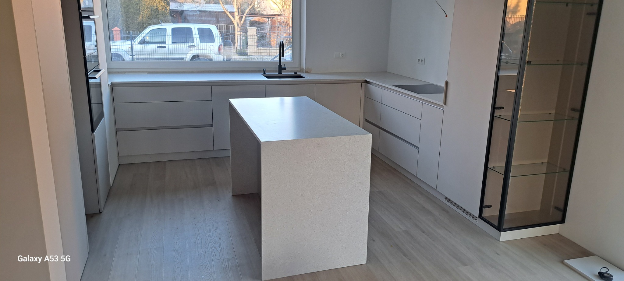 Jasna kuchnia w Zapałowie z wyspą i zabudową meblową w minimalistycznym stylu, widok na okno z zabudową.