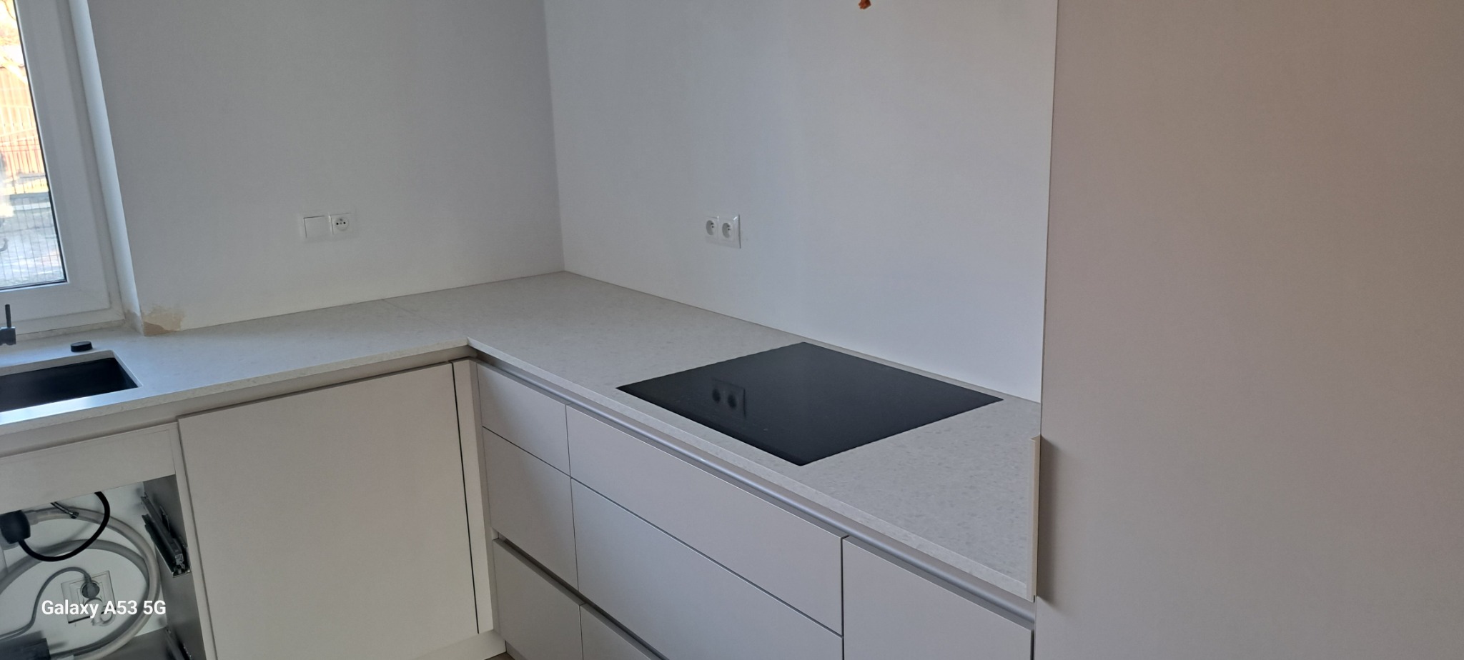 Jasna kuchnia w trakcie wykańczania, widoczny blat z szarego konglomeratu z zamontowaną płytą indukcyjną, białe szafki z szufladami i zlew przy oknie.