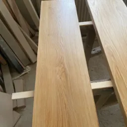 Dwie świeżo polakierowane deski z jasnego drewna dębowego, ułożone na drewnianych kozłach w warsztacie, widoczne naturalne usłojenie drewna.