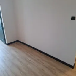 Fragment pokoju po remoncie: świeżo pomalowane ściany w odcieniu złamanej bieli, nowe panele podłogowe w ciepłym odcieniu drewna oraz czarne listwy przypodłogowe. Widoczny czarny włącznik światła...