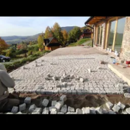 Układanie kostki brukowej na tarasie domu z widokiem na góry, częściowo ułożona nawierzchnia z kostki granitowej, przygotowane kamienie i piasek.