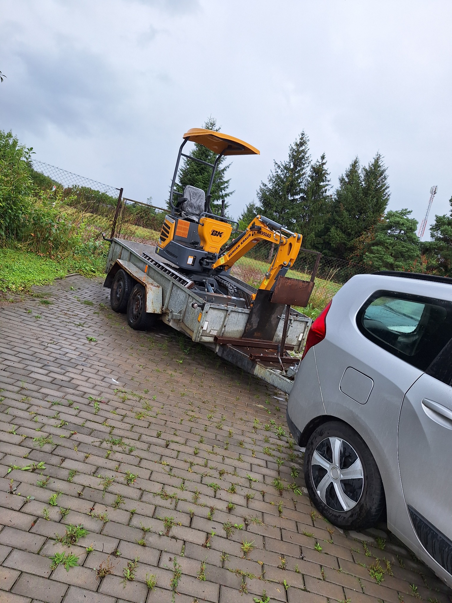 Żółta mini koparka Berger Kraus BK950AS na przyczepie, zaparkowana obok srebrnego samochodu na kostce brukowej z chwastami, w tle ogrodzenie z siatki i drzewa iglaste pod pochmurnym niebem.