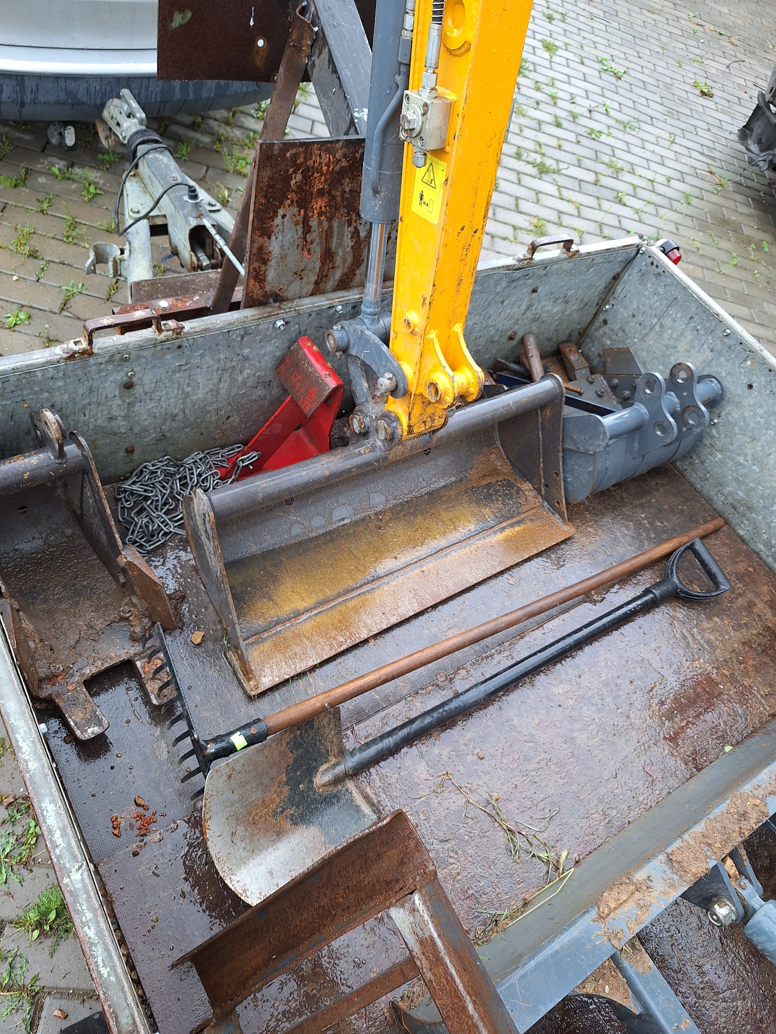 Przyczepa z żółtym ramieniem koparki, łyżkami, łańcuchem, łopatami i grabiami, wszystko pokryte błotem i rdzą.