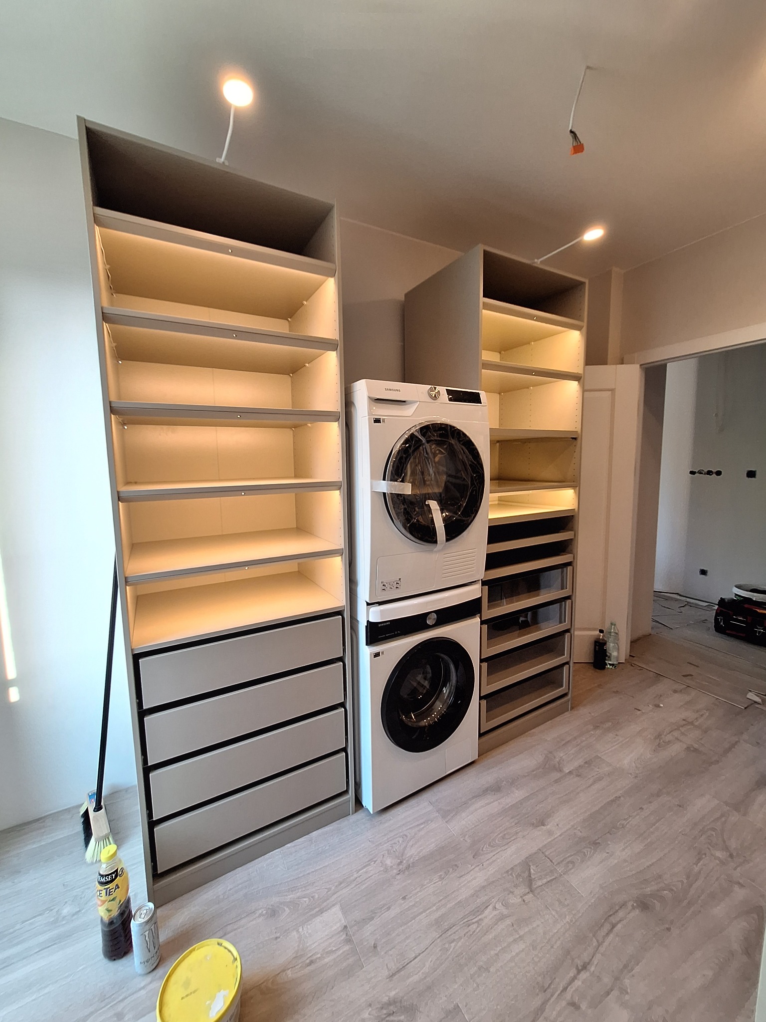 Wnętrze garderoby w trakcie montażu: szafy z szufladami i półkami z podświetleniem LED, pralka i suszarka ustawione w pionie, na podłodze panele, widoczne niedokończone prace wykończeniowe.
