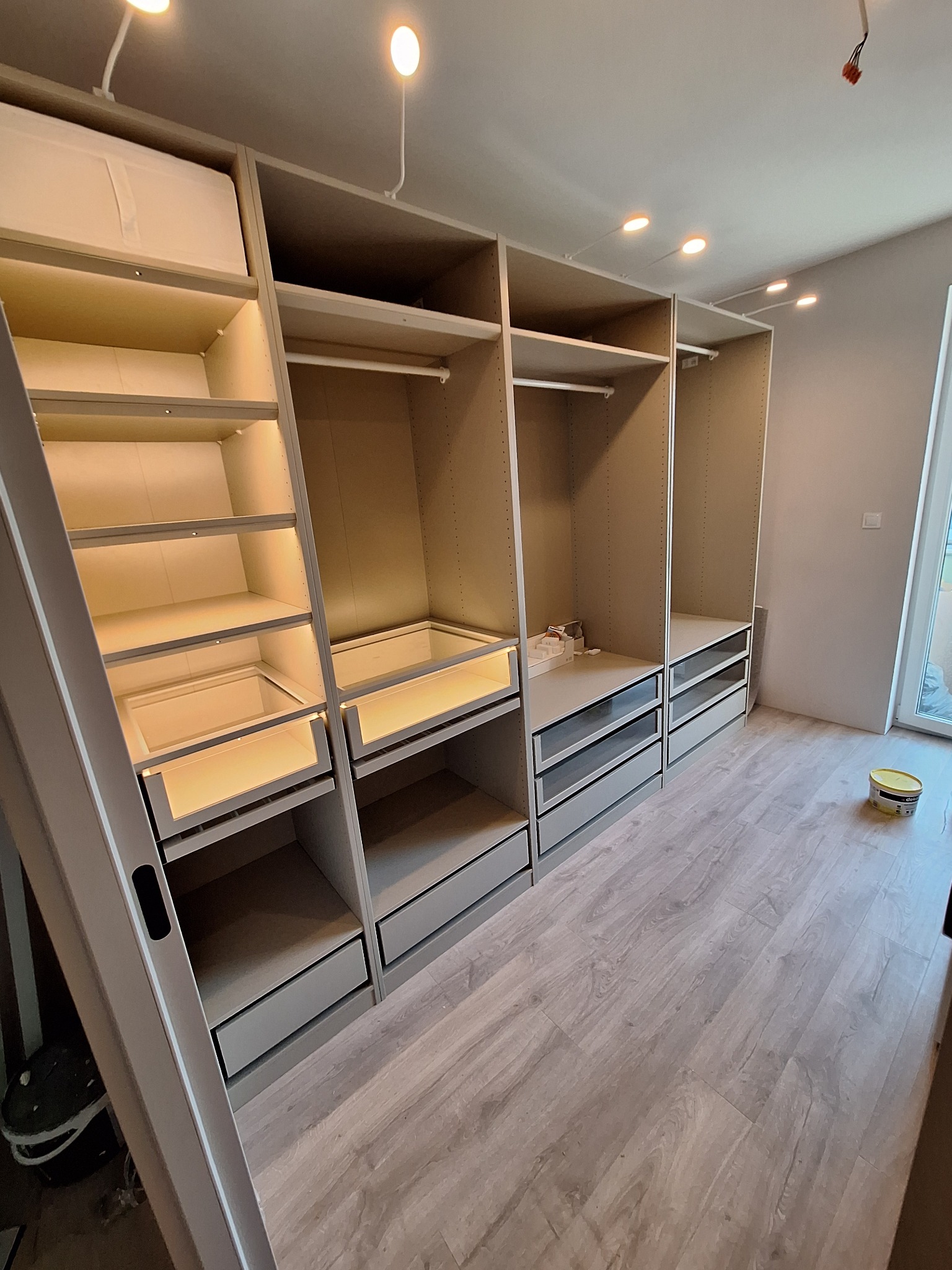 Nowoczesna, szara garderoba z oświetleniem LED wewnątrz szaf, widoczna puszka farby na podłodze z paneli.