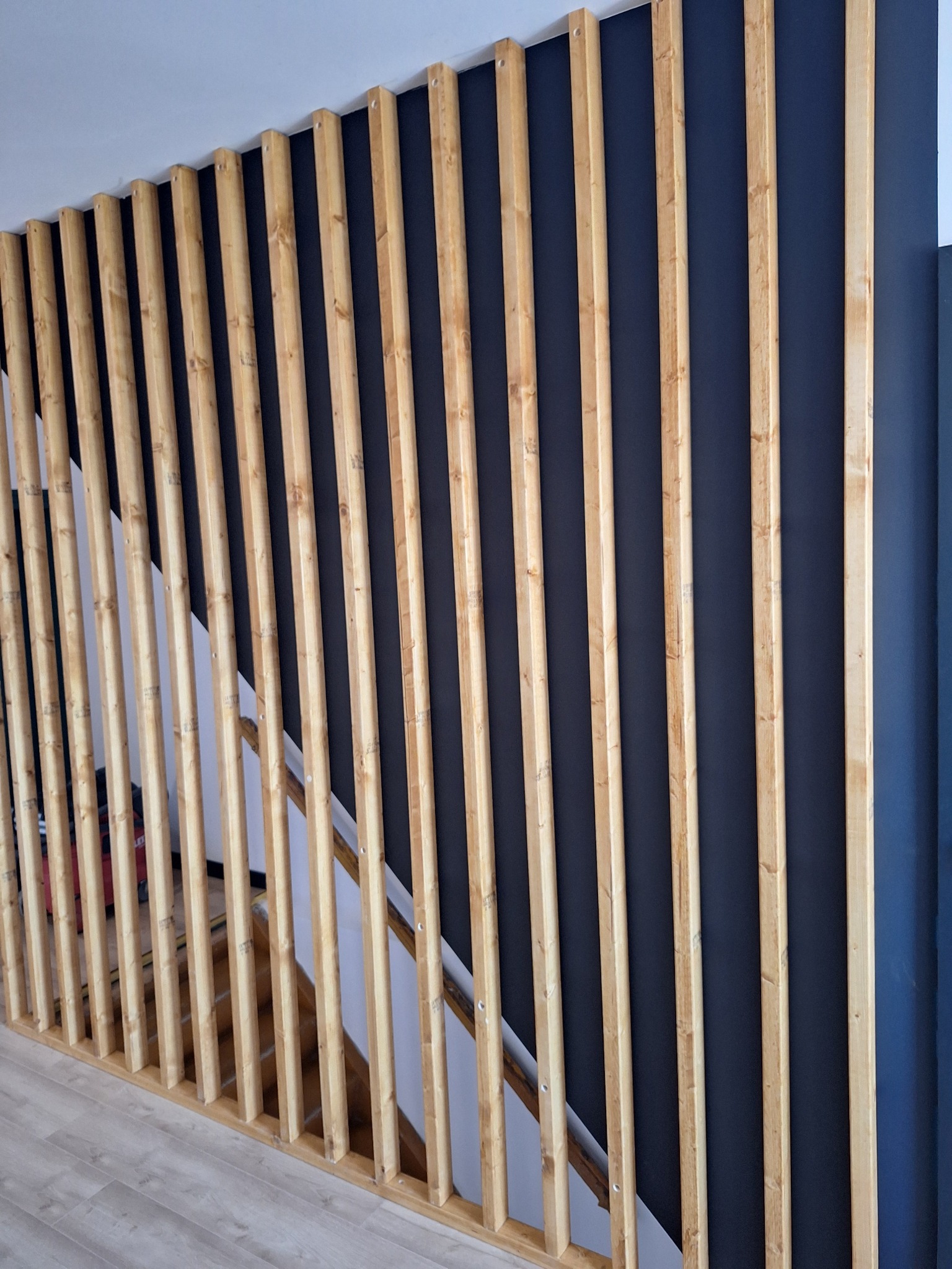 Nowoczesna przegroda schodowa z drewnianych listew na tle granatowej ściany, widok z dołu na schody.