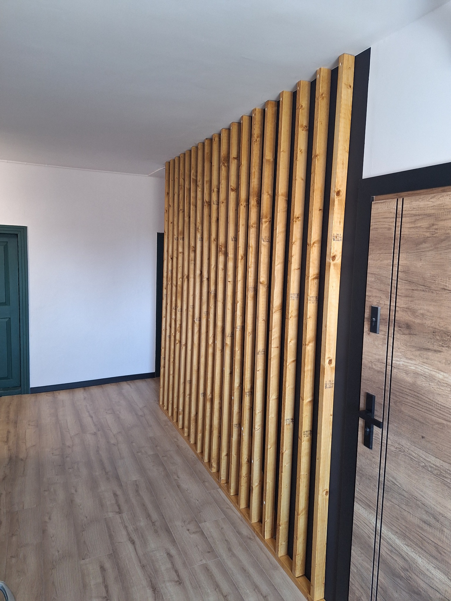 Drewniane lamele tworzące ściankę działową, zamontowane pionowo, z widoczną strukturą drewna i czarnymi elementami wykończeniowymi, na podłodze panele imitujące drewno, w tle biała ściana i zielone...