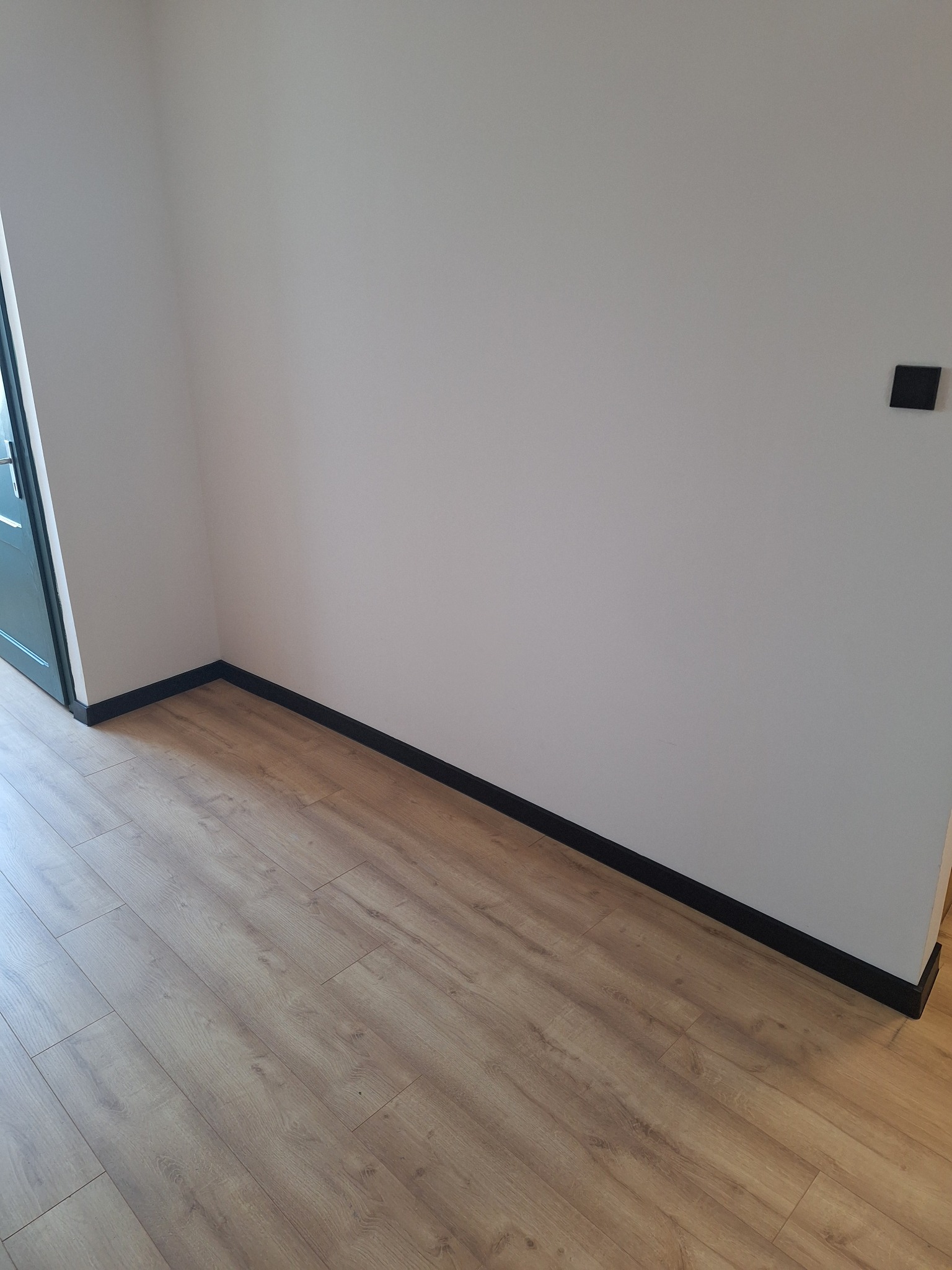 Fragment pokoju po remoncie: świeżo pomalowane ściany w odcieniu złamanej bieli, nowe panele podłogowe w ciepłym odcieniu drewna oraz czarne listwy przypodłogowe. Widoczny czarny włącznik światła...
