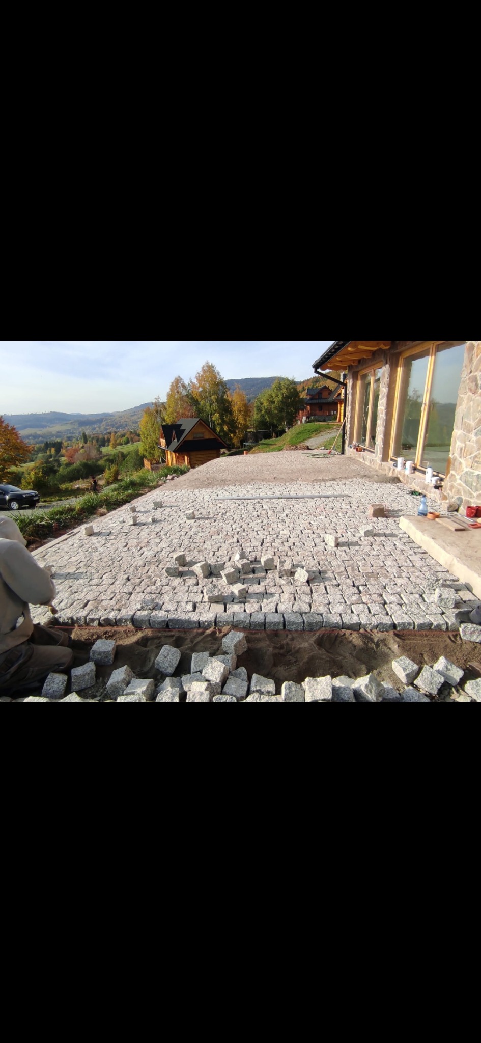 Układanie kostki brukowej na tarasie domu z widokiem na góry, częściowo ułożona nawierzchnia z kostki granitowej, przygotowane kamienie i piasek.