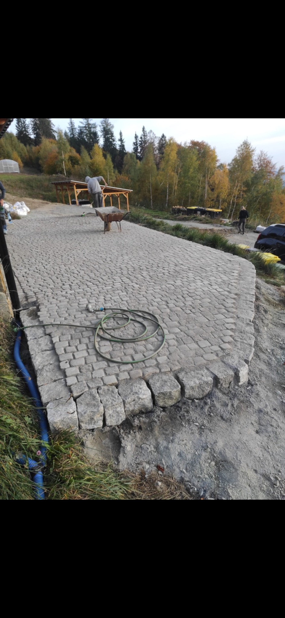 Układanie kostki brukowej na posesji, widoczny fragment ułożonej nawierzchni z kostki, zwinięty wąż ogrodowy i pracujące osoby w tle.
