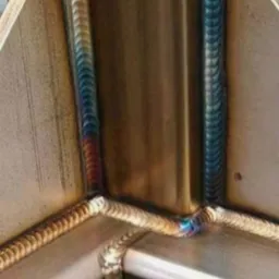 Szczegółowe zbliżenie na precyzyjne spawy łączące metalowe elementy pod kątem prostym, z widocznymi śladami ciepła w postaci tęczowych przebarwień.