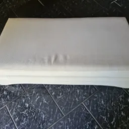 Pufa tapicerowana jasną tkaniną o drobnej strukturze, leżąca na podłodze wyłożonej czarno-białymi płytkami imitującymi marmur.