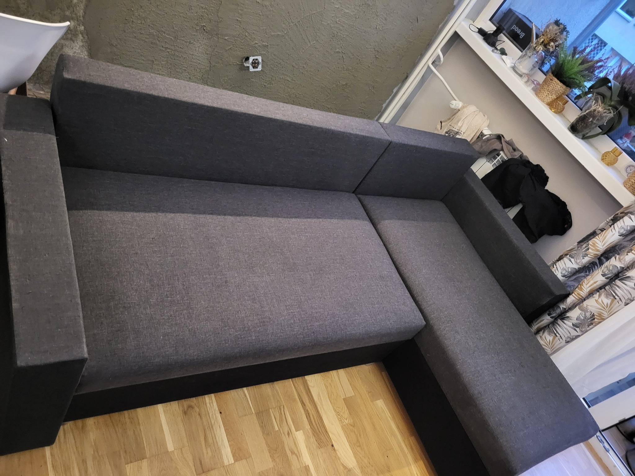 Szara, narożna sofa z tapicerką o wyraźnej fakturze, widziana z góry, na drewnianej podłodze; za sofą widoczny parapet z dekoracjami.