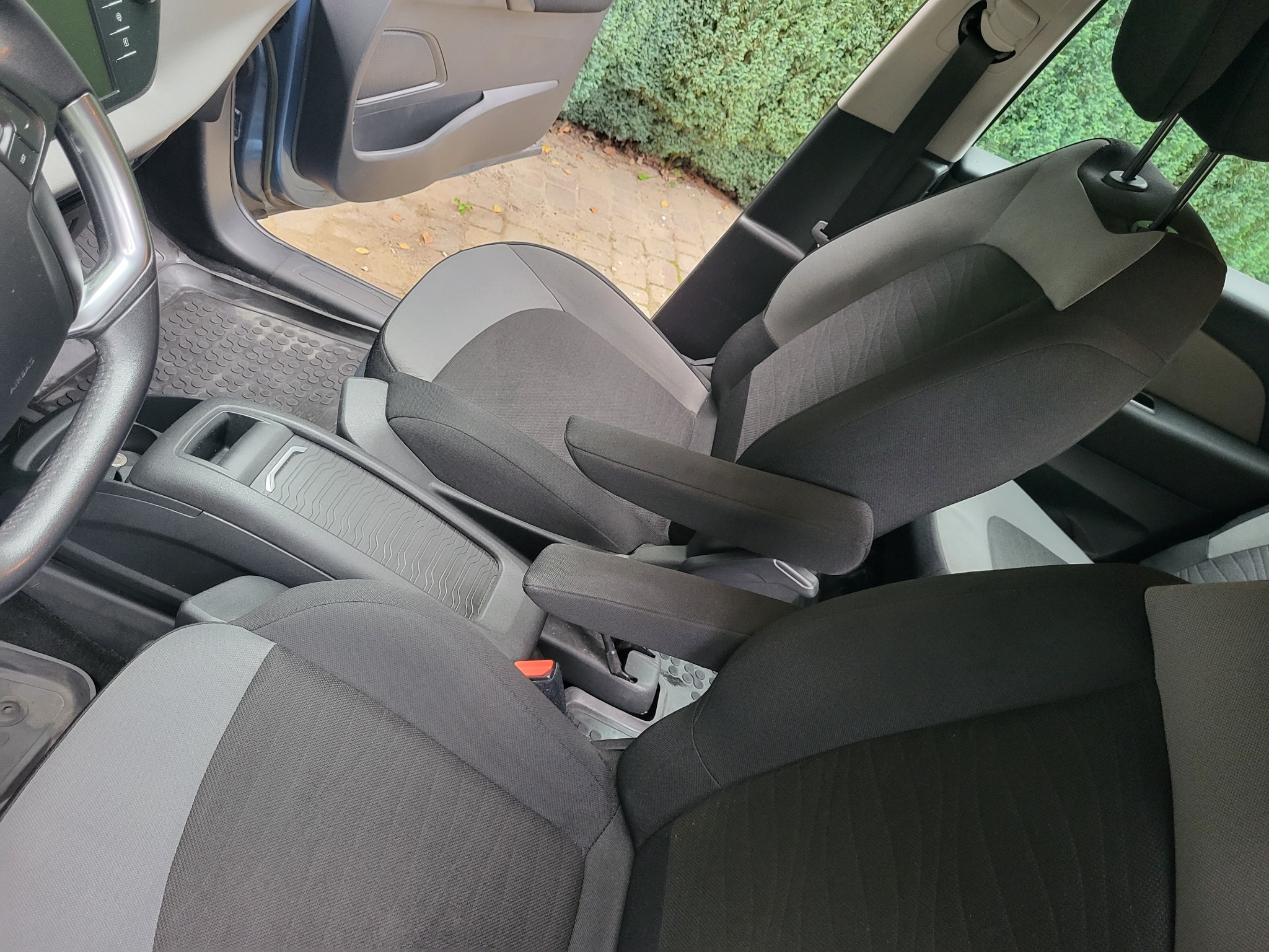 Wnętrze samochodu z widocznymi, odnowionymi fotelami tapicerowanymi w odcieniach szarości i czerni, z podłokietnikami, z otwartymi drzwiami i widokiem na zielony żywopłot na zewnątrz.