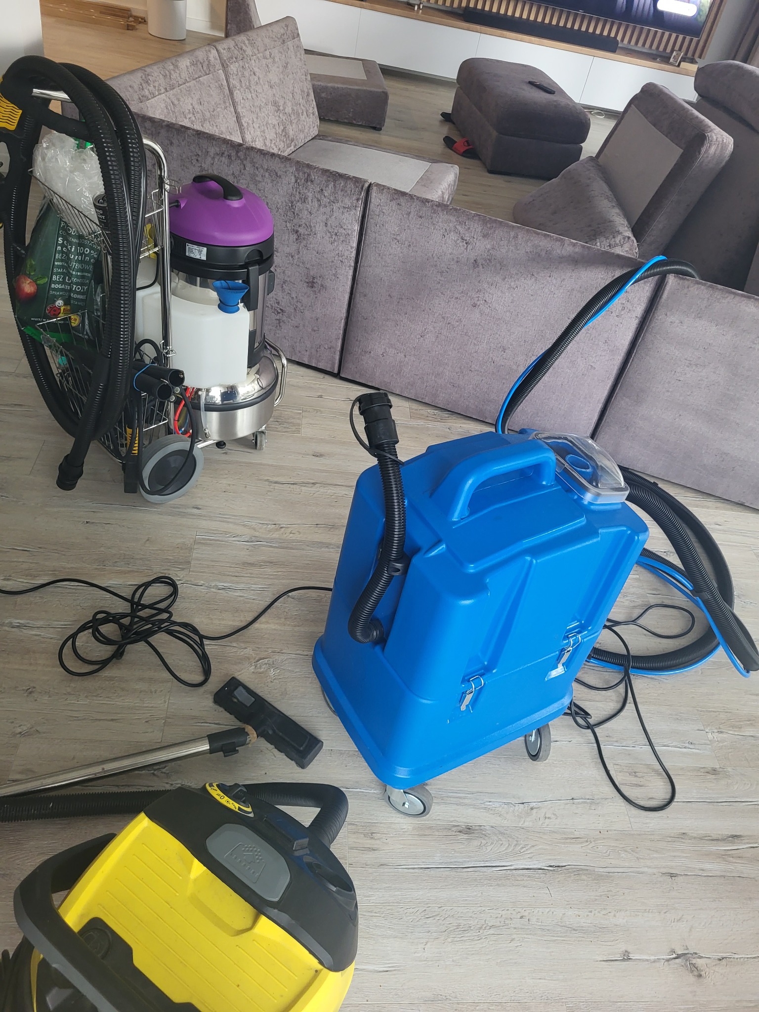 Profesjonalne urządzenia do czyszczenia tapicerki, w tym odkurzacze piorące i akcesoria, rozstawione na drewnianej podłodze przed szarą sofą modułową.