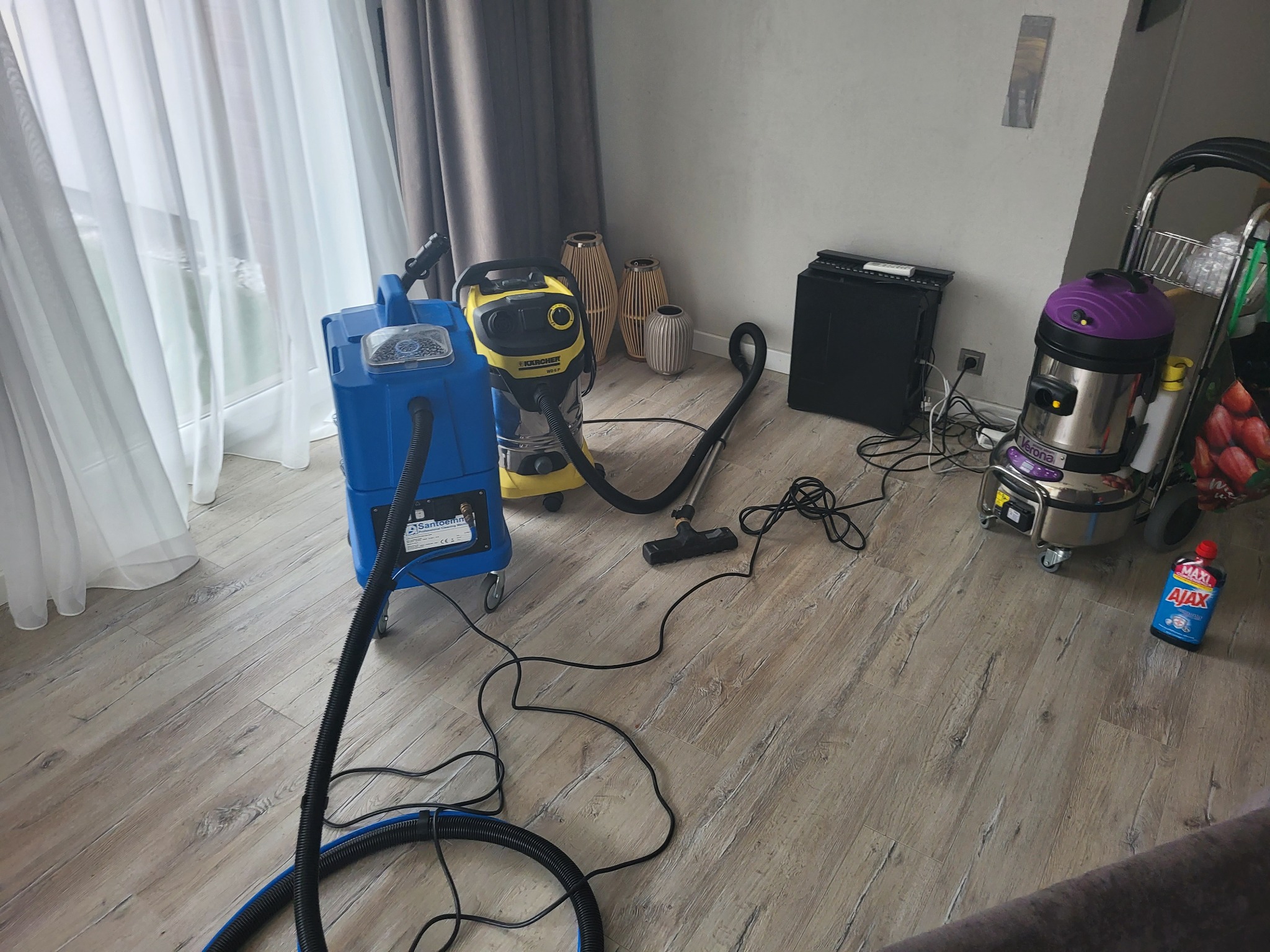 Sprzęt do prania dywanów i tapicerki w Łodzi: niebieski odkurzacz piorący Santoemma, żółty Karcher WD 6 P oraz Verona na wózku, widoczne na drewnianej podłodze, obok okna z białymi zasłonami.