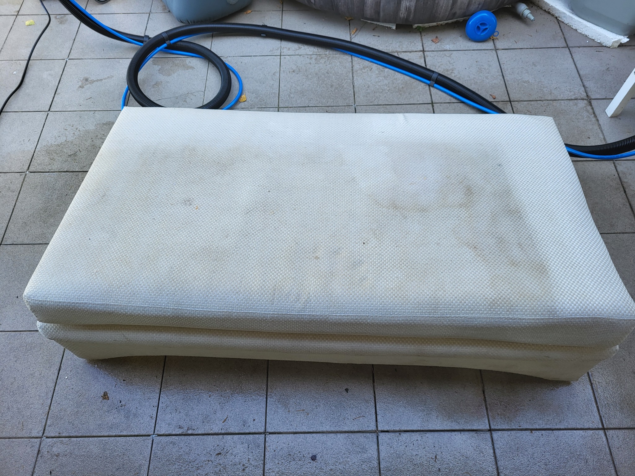 Tapicerowany, kremowy puf z widocznymi plamami, leżący na szarej, kwadratowej płytce, obok czarnego węża z niebieskimi elementami.