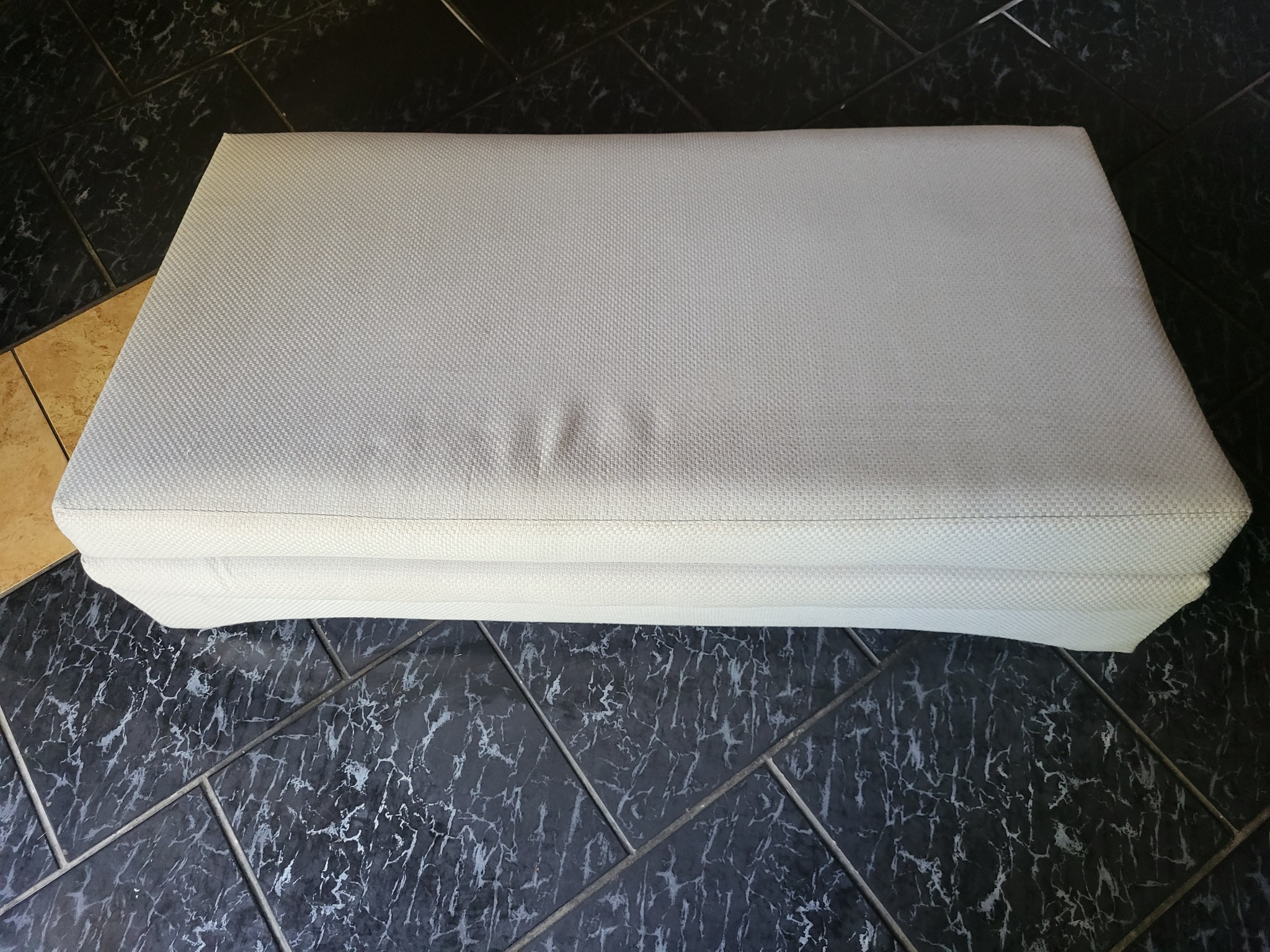 Pufa tapicerowana jasną tkaniną o drobnej strukturze, leżąca na podłodze wyłożonej czarno-białymi płytkami imitującymi marmur.