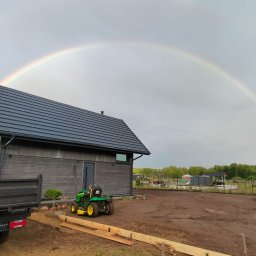 Dachowiec - Nowoczesny dom z ciemną, skośną dachówką w promieniach tęczy. Widoczny fragment ciężarówki i traktorka ogrodowego. Ujęcie z ziemi.
