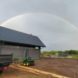 Nowoczesny dom z ciemną, skośną dachówką w promieniach tęczy. Widoczny fragment ciężarówki i traktorka ogrodowego. Ujęcie z ziemi.
