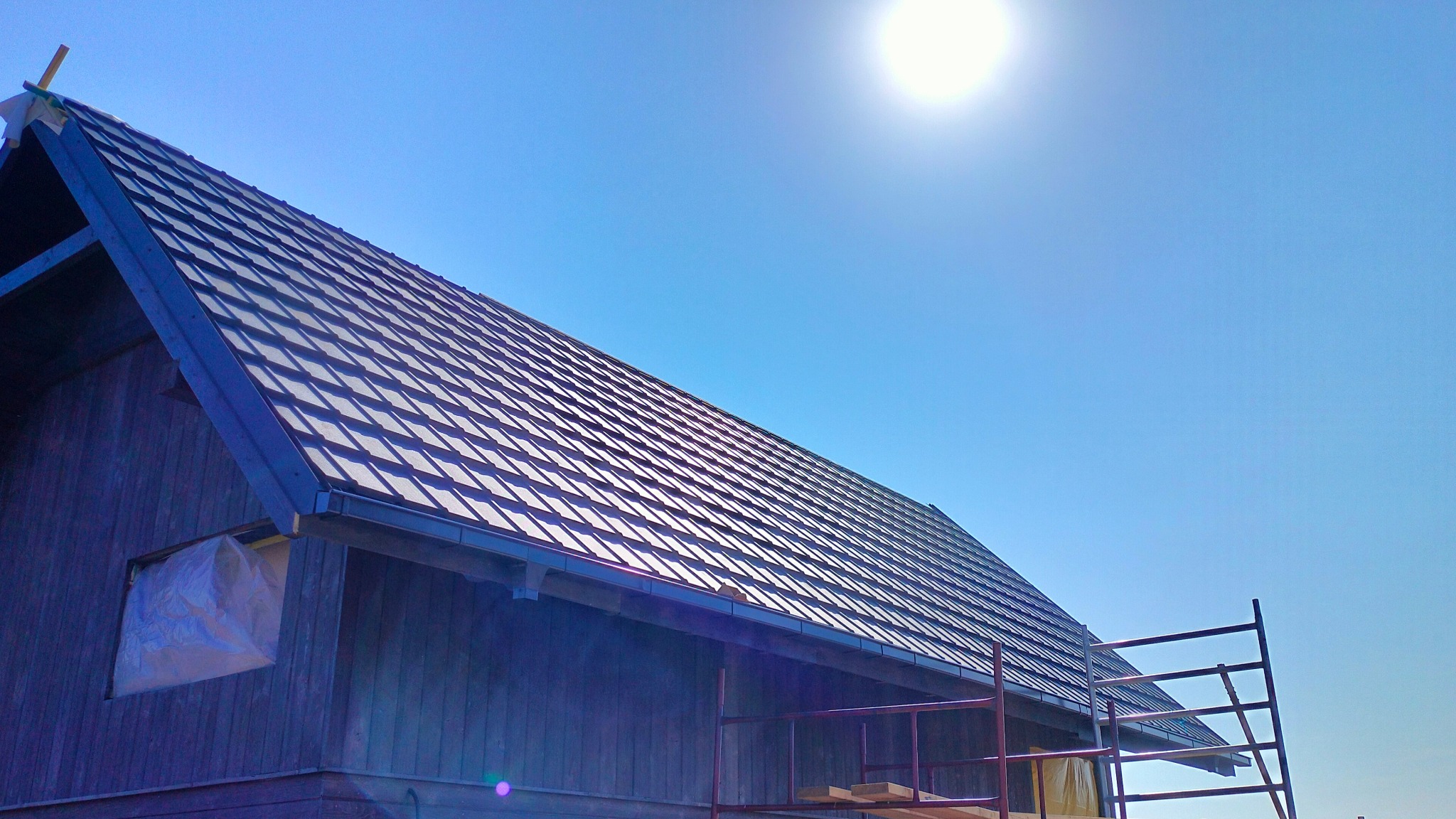 Nowy, ciemny dach z blachy trapezowej na drewnianym budynku w trakcie budowy, widoczne rusztowanie i zakryte okno, jasne, słoneczne niebo.