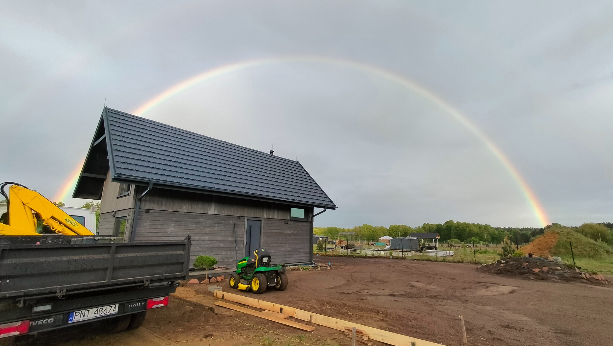 Nowoczesny dom z ciemną, skośną dachówką w promieniach tęczy. Widoczny fragment ciężarówki i traktorka ogrodowego. Ujęcie z ziemi.
