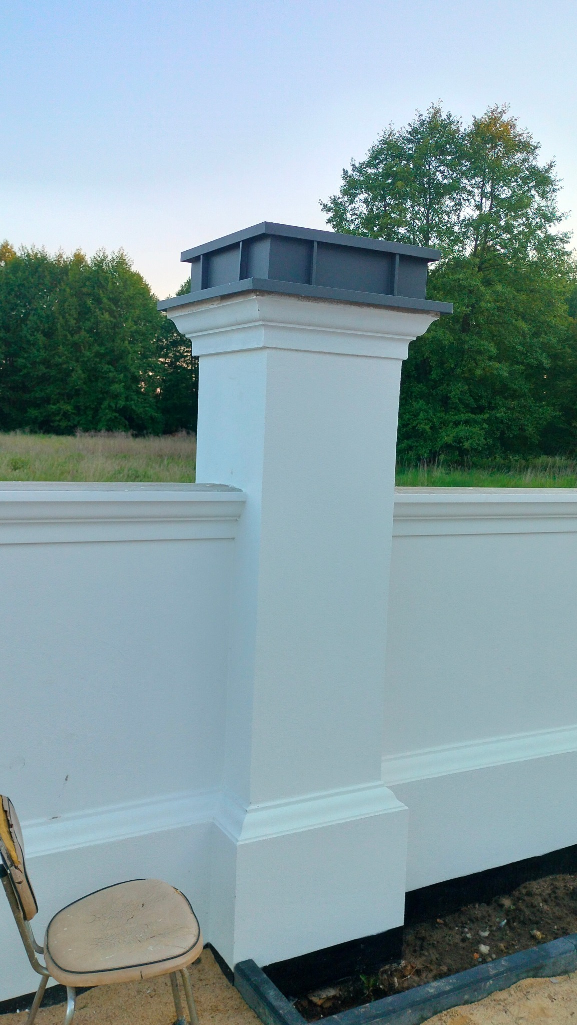 Białe, otynkowane ogrodzenie z filarami zwieńczonymi szarymi elementami dekoracyjnymi, na tle zieleni drzew i nieba. Widoczny fragment krzesła.