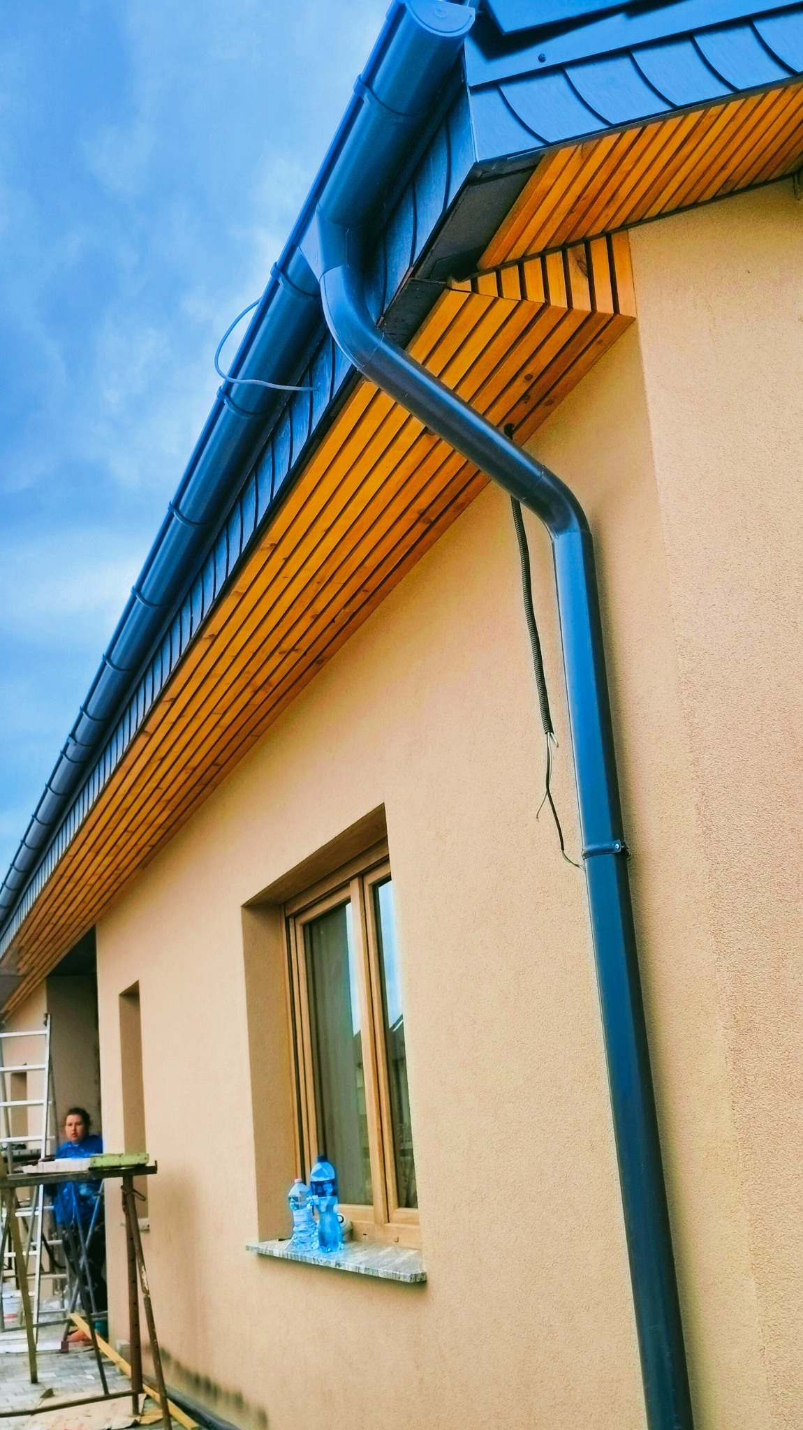 Zamontowana, ciemna rynna spustowa na elewacji budynku z drewnianą podbitką. W tle pracownik na rusztowaniu.