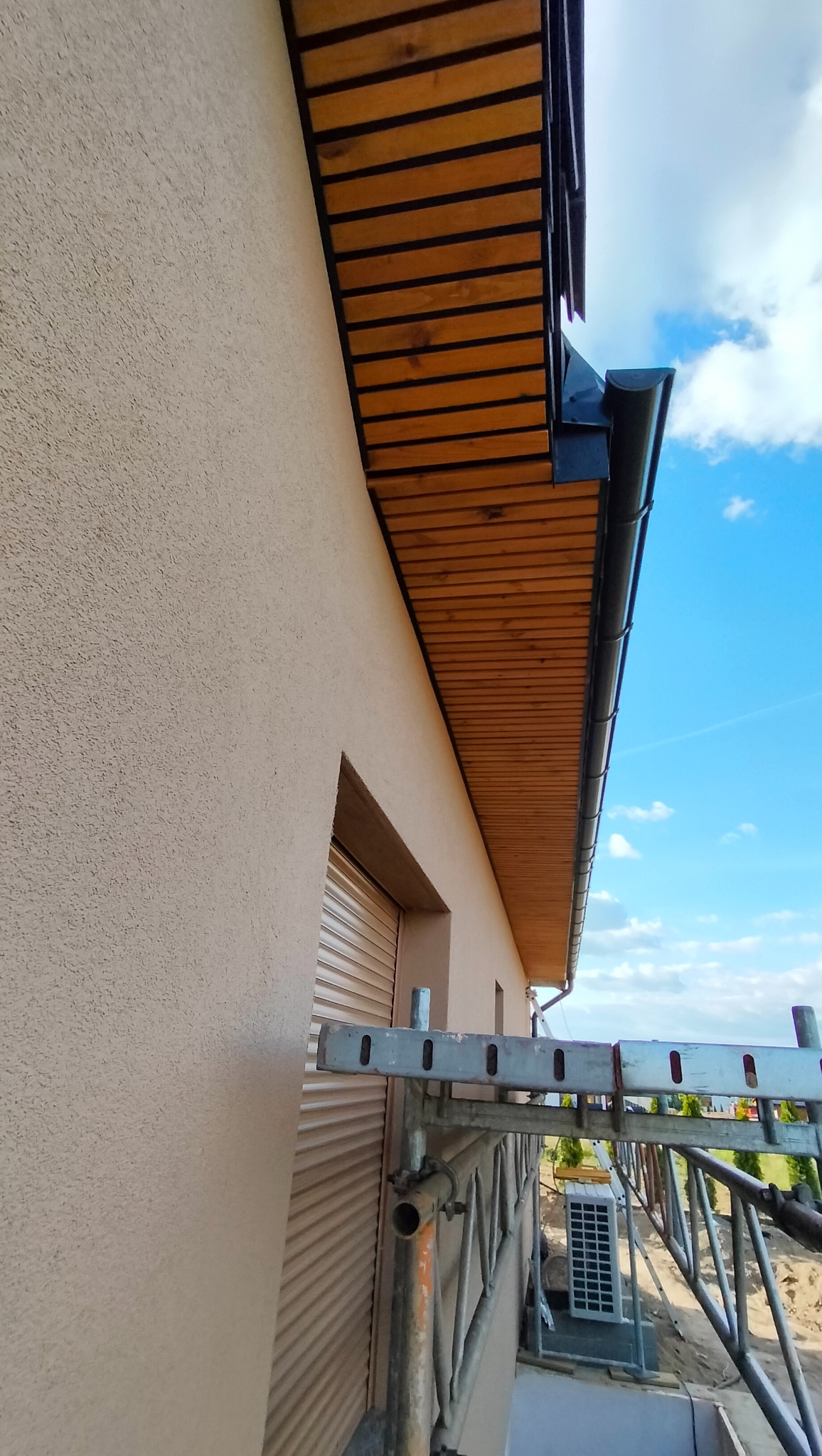 Elewacja domu z zamontowanym systemem rynnowym i drewnianą podbitką, widok z rusztowania. Błękitne niebo w tle, jasna elewacja.