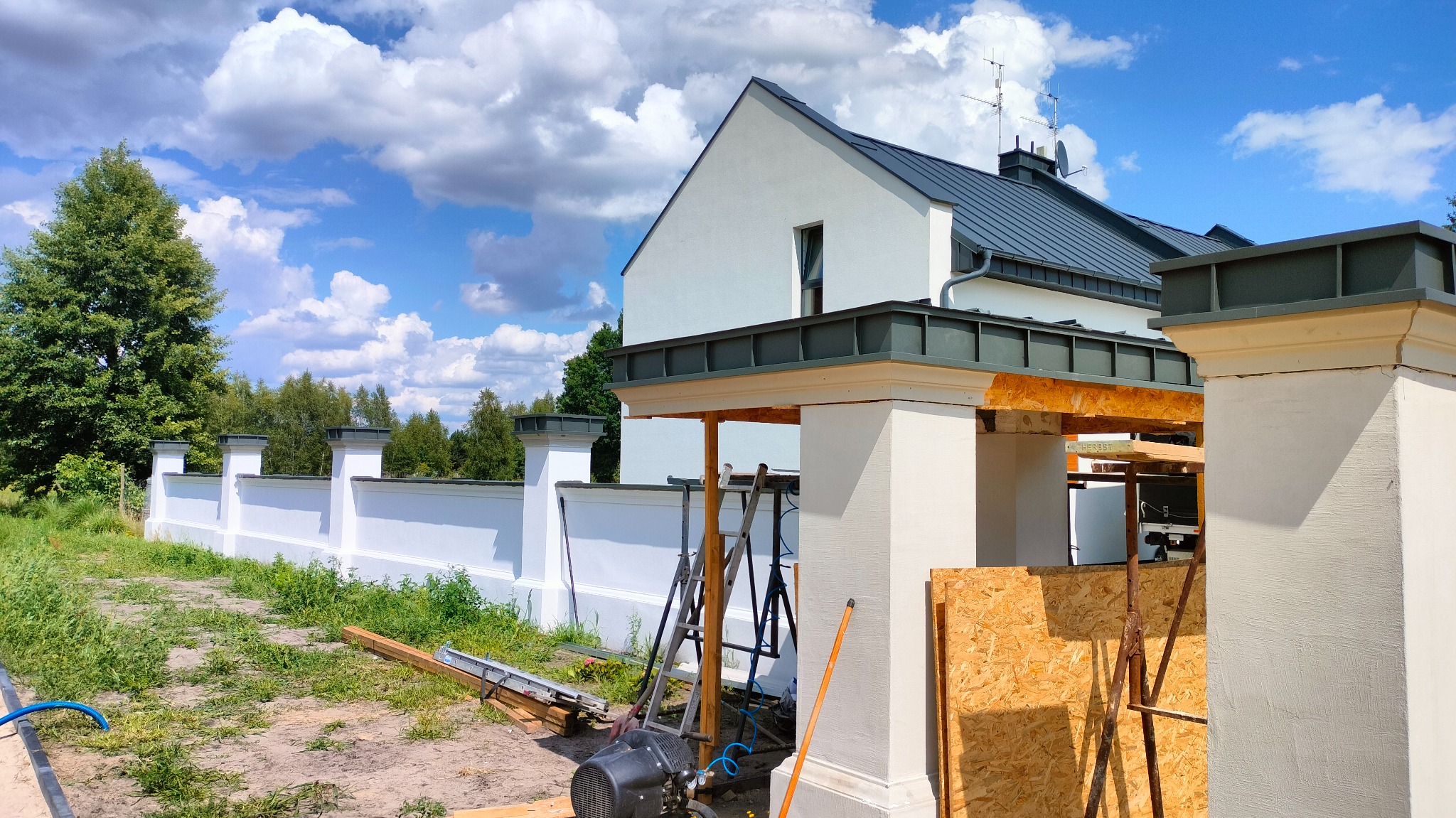 Biały dom z szarym dachem w trakcie budowy, widoczne rusztowania i narzędzia. Elementy obróbki blacharskiej na dachu. Białe ogrodzenie w tle.