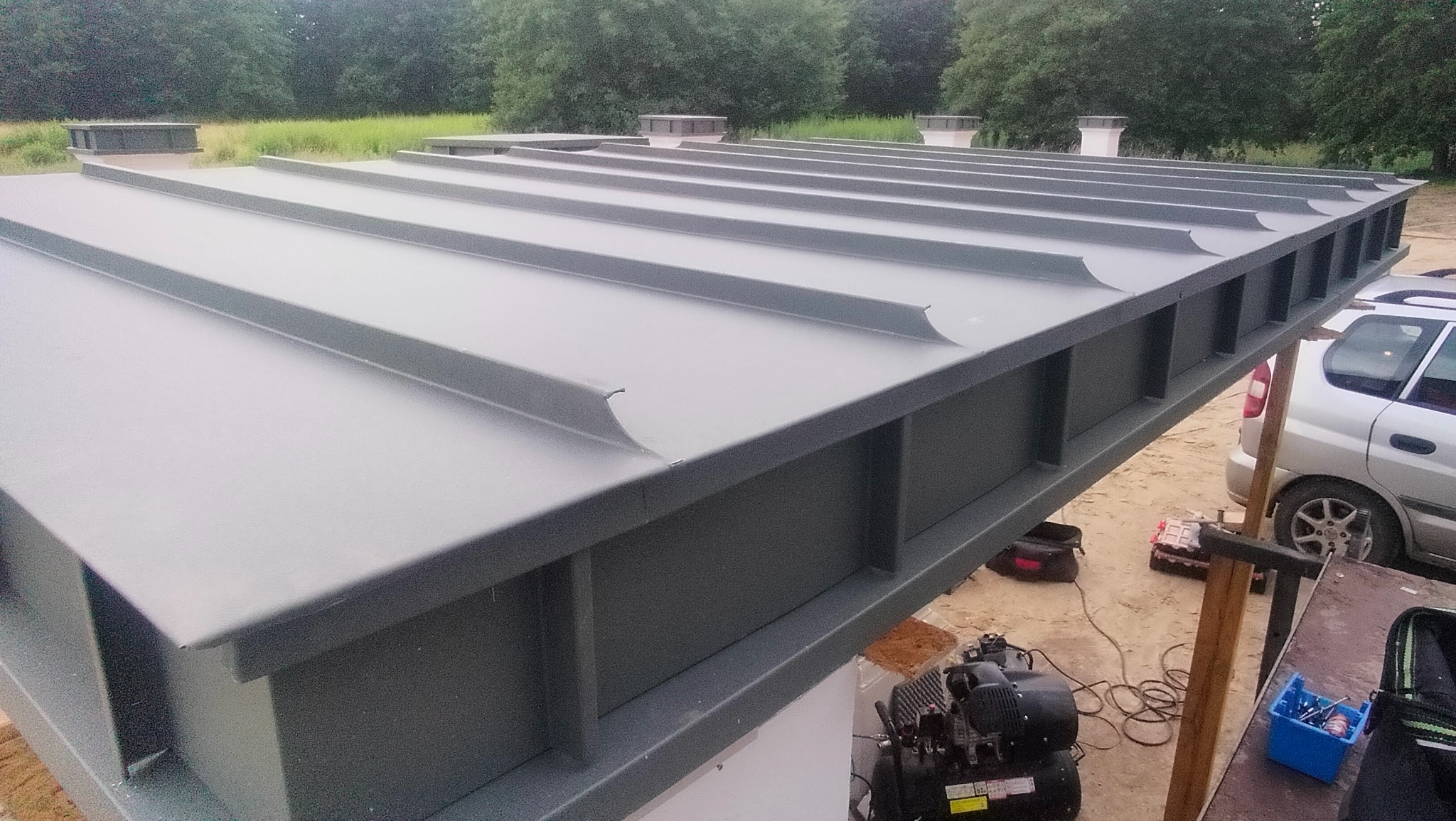 Szary dach z blachy trapezowej z widocznymi elementami konstrukcyjnymi, na tle zieleni i częściowo widocznym samochodem. Widoczne narzędzia i kompresor.