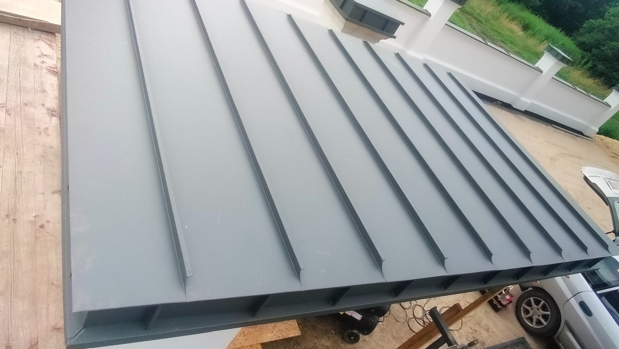 Szary dach z blachy trapezowej z widocznymi łączeniami, sfotografowany z góry pod kątem. W tle białe elementy architektoniczne i zieleń. Częściowo widoczny samochód.