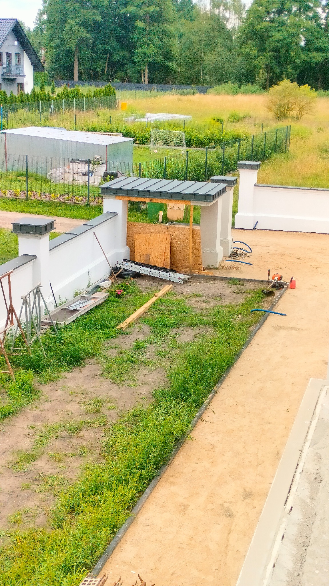 Wysoki kąt: budowa zadaszenia nad wejściem, widoczne rusztowania, drabina i narzędzia. W tle dom i zieleń. Prace w toku, etap budowy zadaszenia.