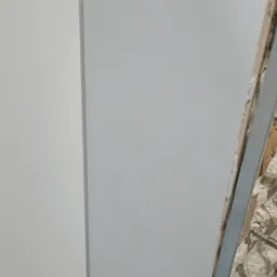 Fragment ściany z zamontowaną białą płytą, widoczne niedoskonałości ściany przed obłożeniem i metalowy profil mocujący.