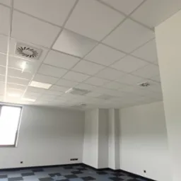 Białe panele sufitowe w biurze z kratką wentylacyjną, wzorzysta podłoga w szaro-niebieską szachownicę, widok na jasne okno.