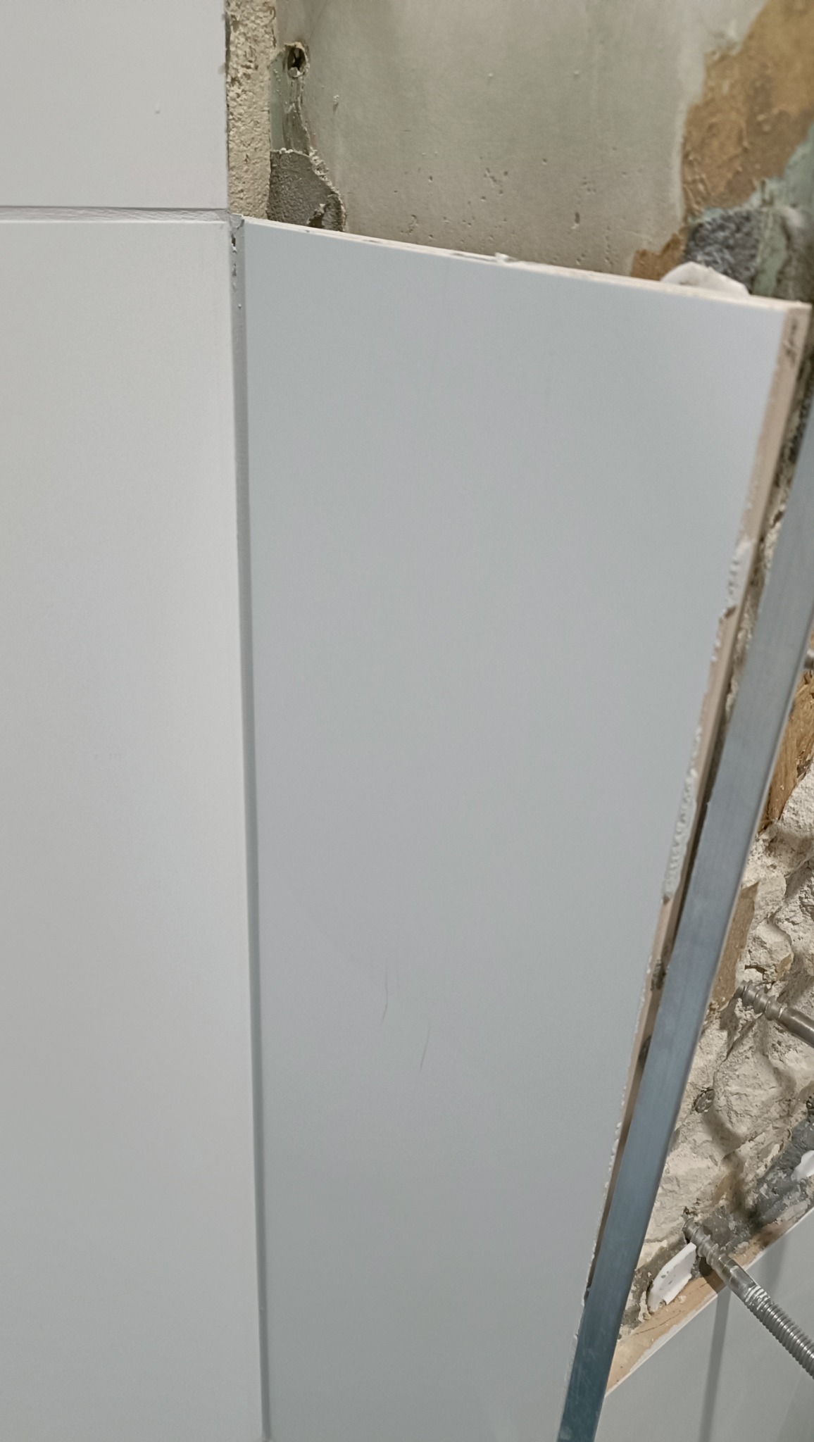 Fragment ściany z zamontowaną białą płytą, widoczne niedoskonałości ściany przed obłożeniem i metalowy profil mocujący.