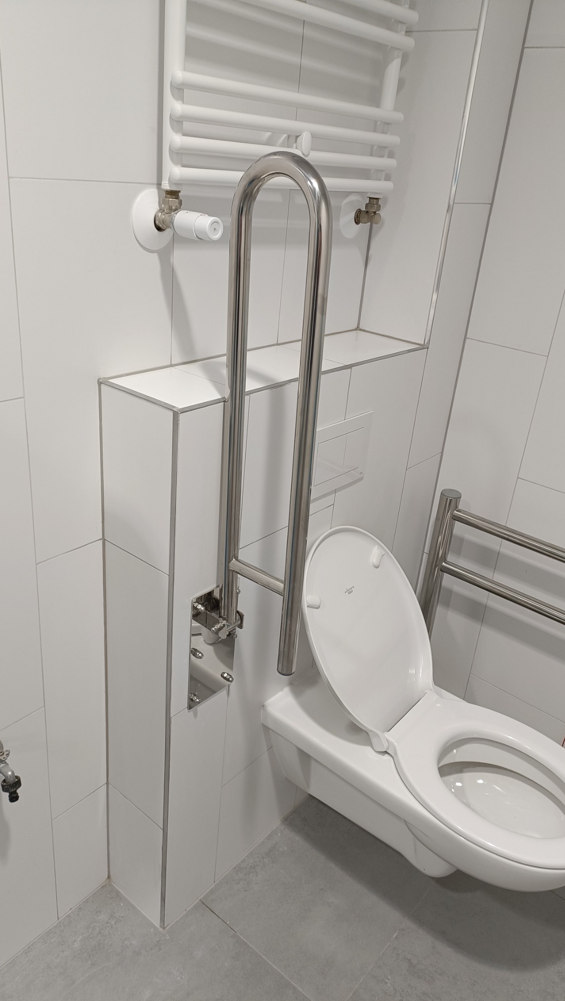 Biała, podwieszana toaleta z otwartą klapą i zamontowanym chromowanym uchwytem dla osób niepełnosprawnych, na tle białych płytek i grzejnika drabinkowego.