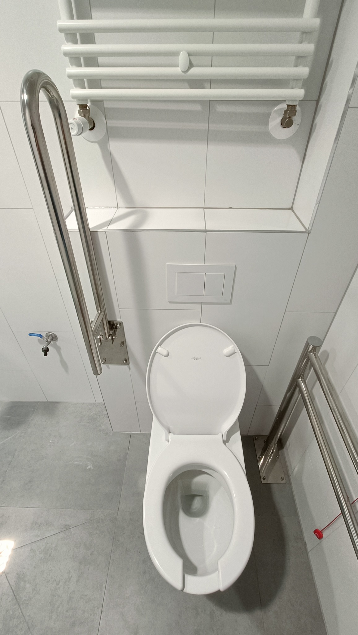 Biała toaleta z otwartą klapą w nowoczesnej łazience, wyposażona w chromowany uchwyt dla osób z niepełnosprawnościami i grzejnik drabinkowy na ścianie wyłożonej białymi płytkami.