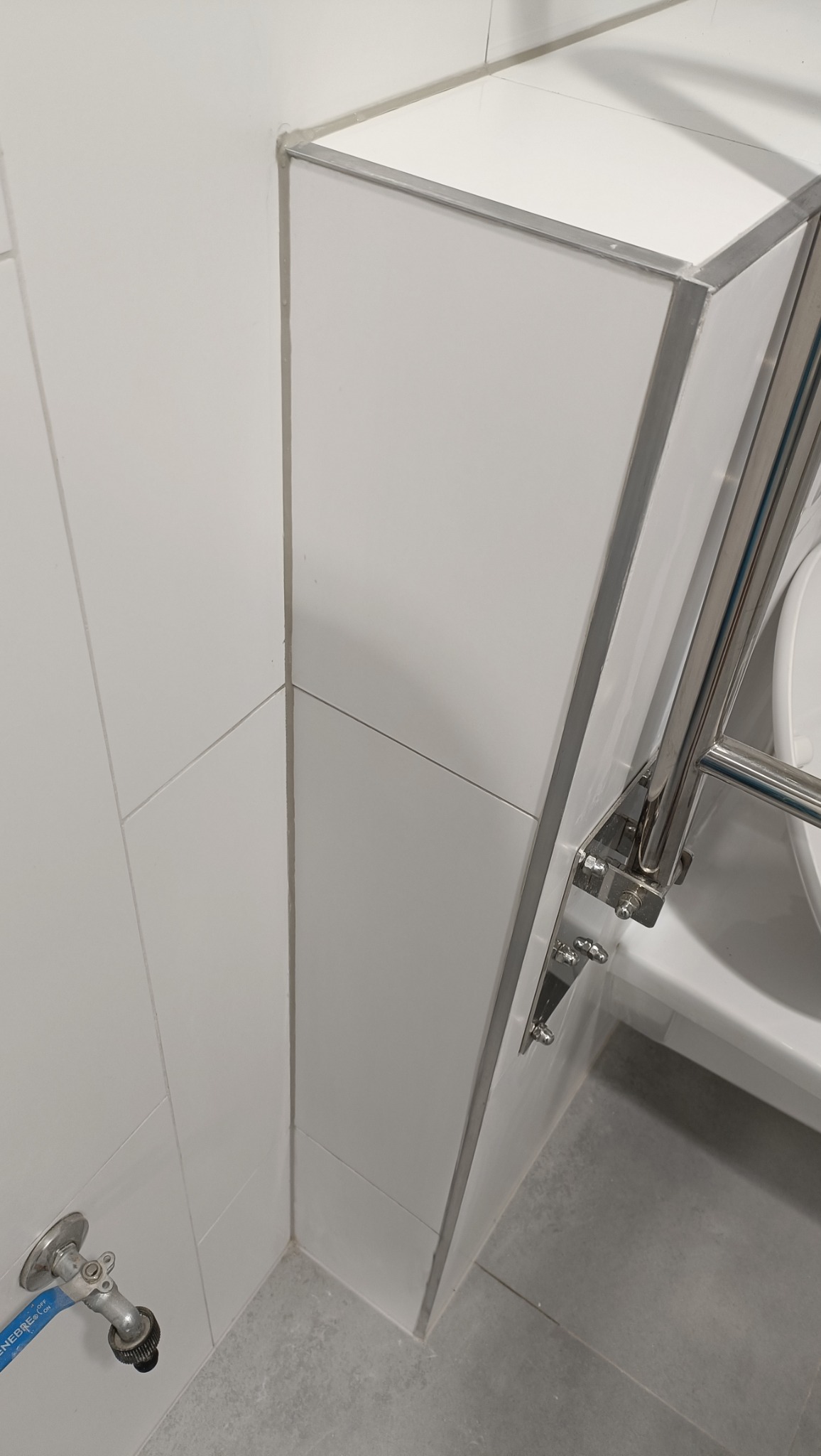 Detal wykończenia łazienki: białe, ukośne płytki ścienne z metalowymi listwami, fragment grzejnika drabinkowego i zawór wody przy podłodze.