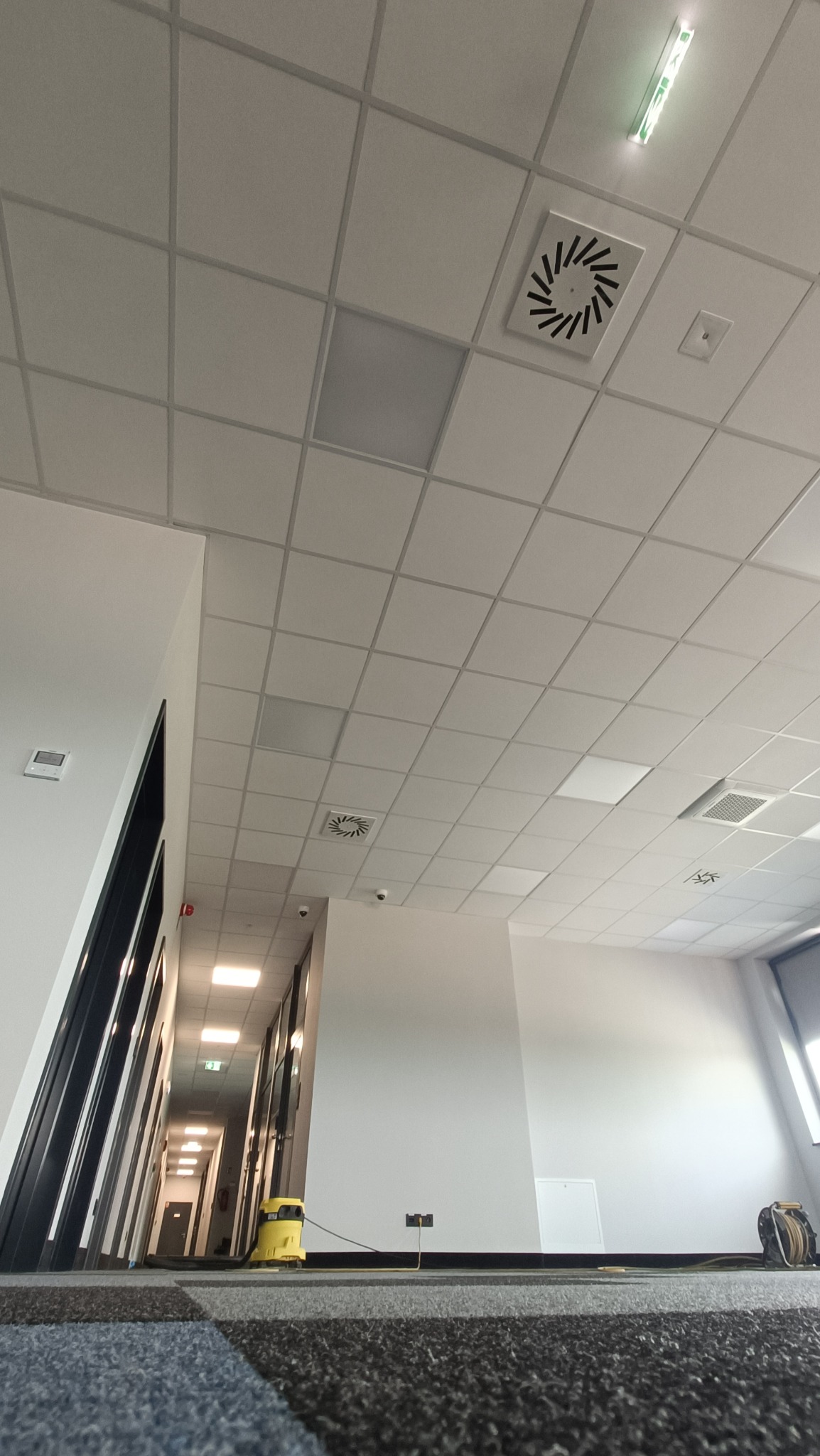 Widok z dołu na sufit podwieszany w biurze, z kratką wentylacyjną i lampą fluorescencyjną, z widocznym korytarzem i sprzętem do czyszczenia podłóg w tle.