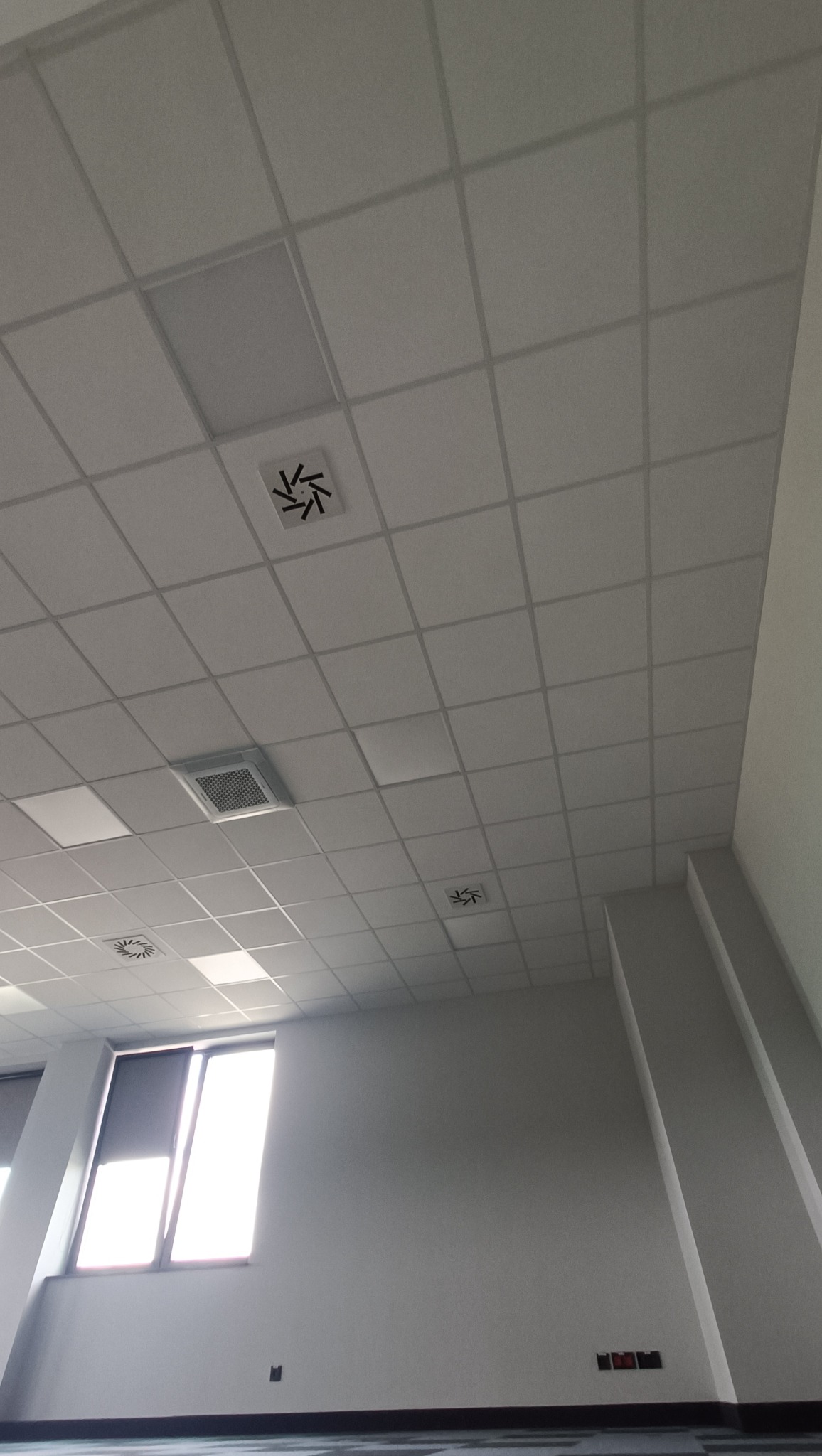 Kasetonowy sufit podwieszany w biurze z kratkami wentylacyjnymi i oknem z roletą.