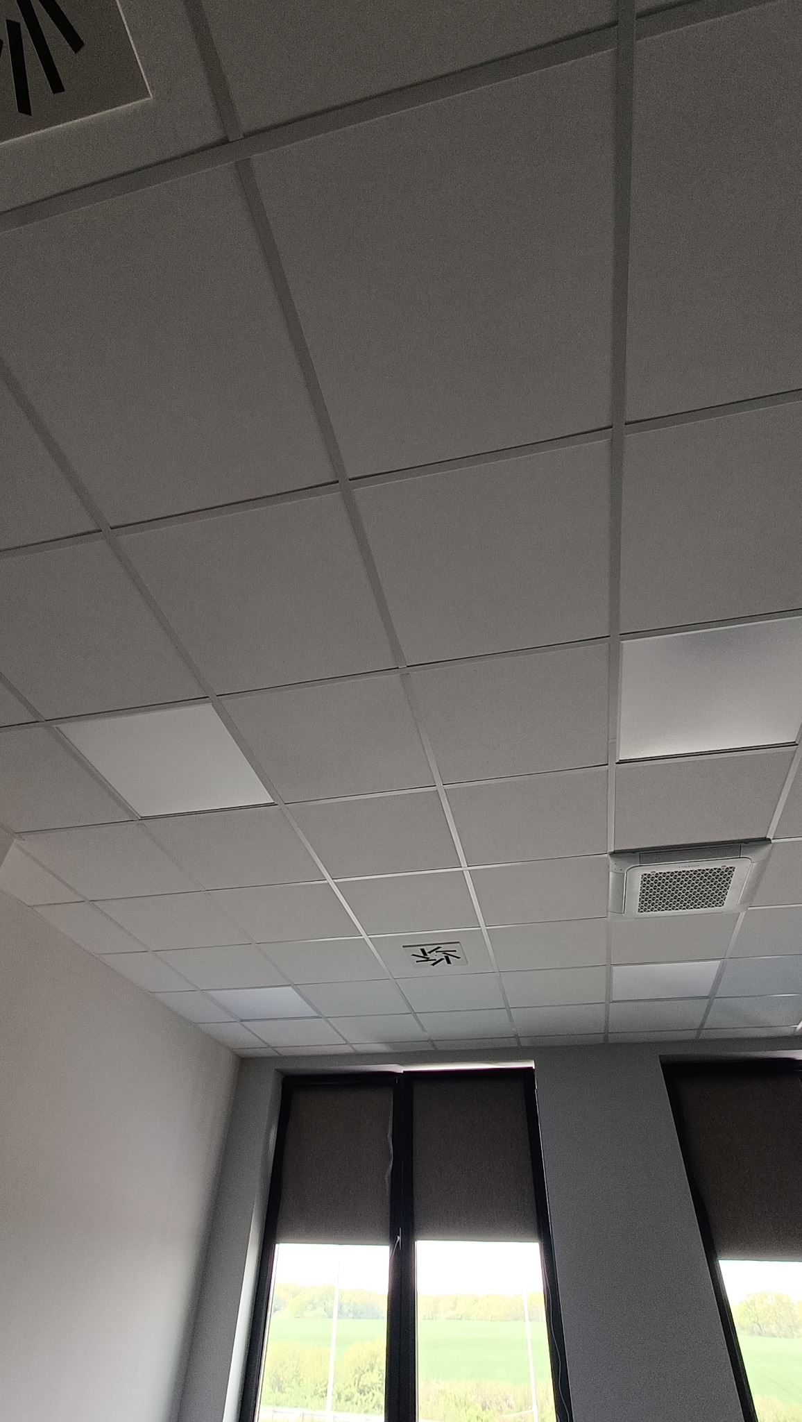 Sufit kasetonowy w biurze z kratką wentylacyjną, widok z dołu na okna z zasłoniętymi roletami i zielenią na zewnątrz.