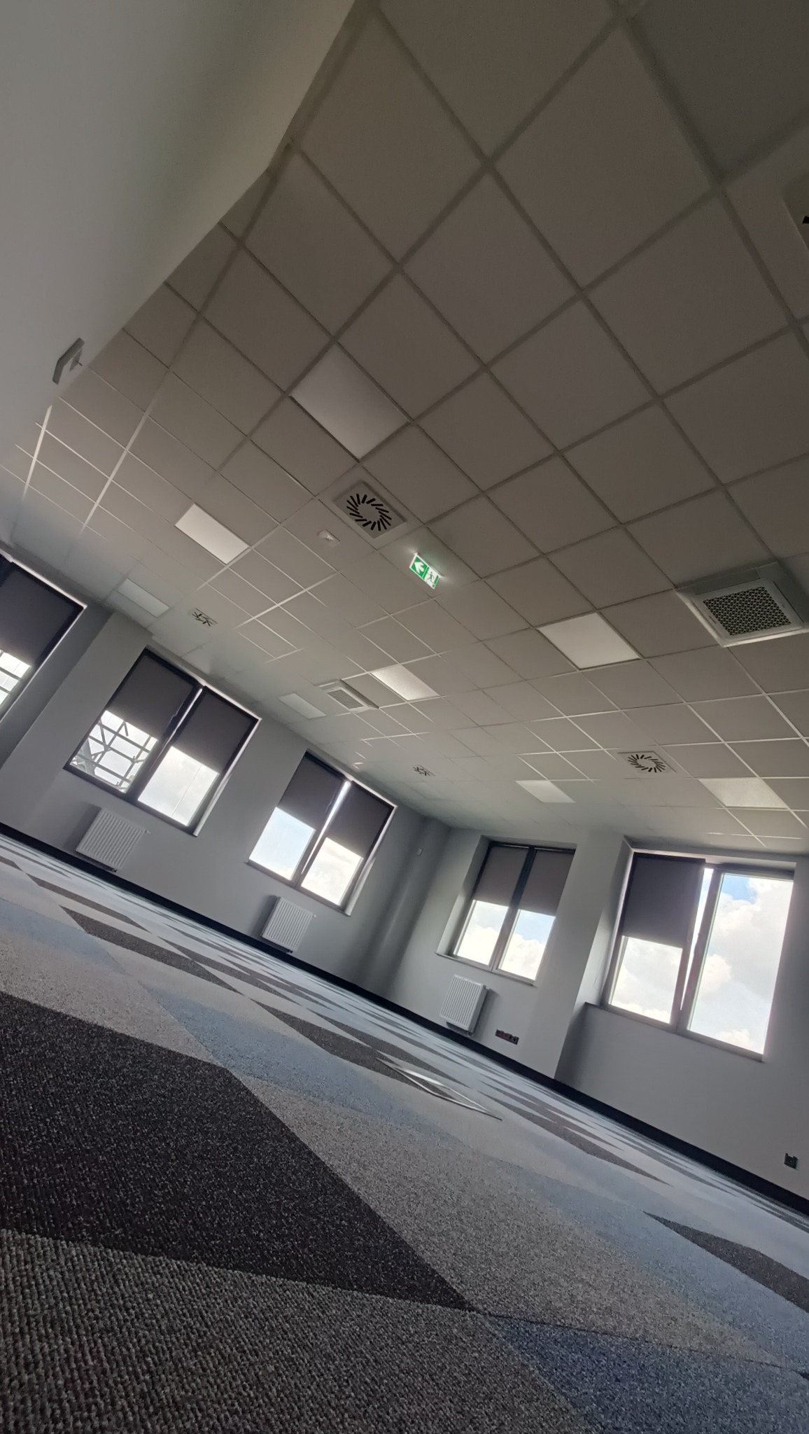 Wnętrze biura z szarą wykładziną w geometryczne wzory, białym sufitem podwieszanym z kratownicą, oknami z roletami i grzejnikami.